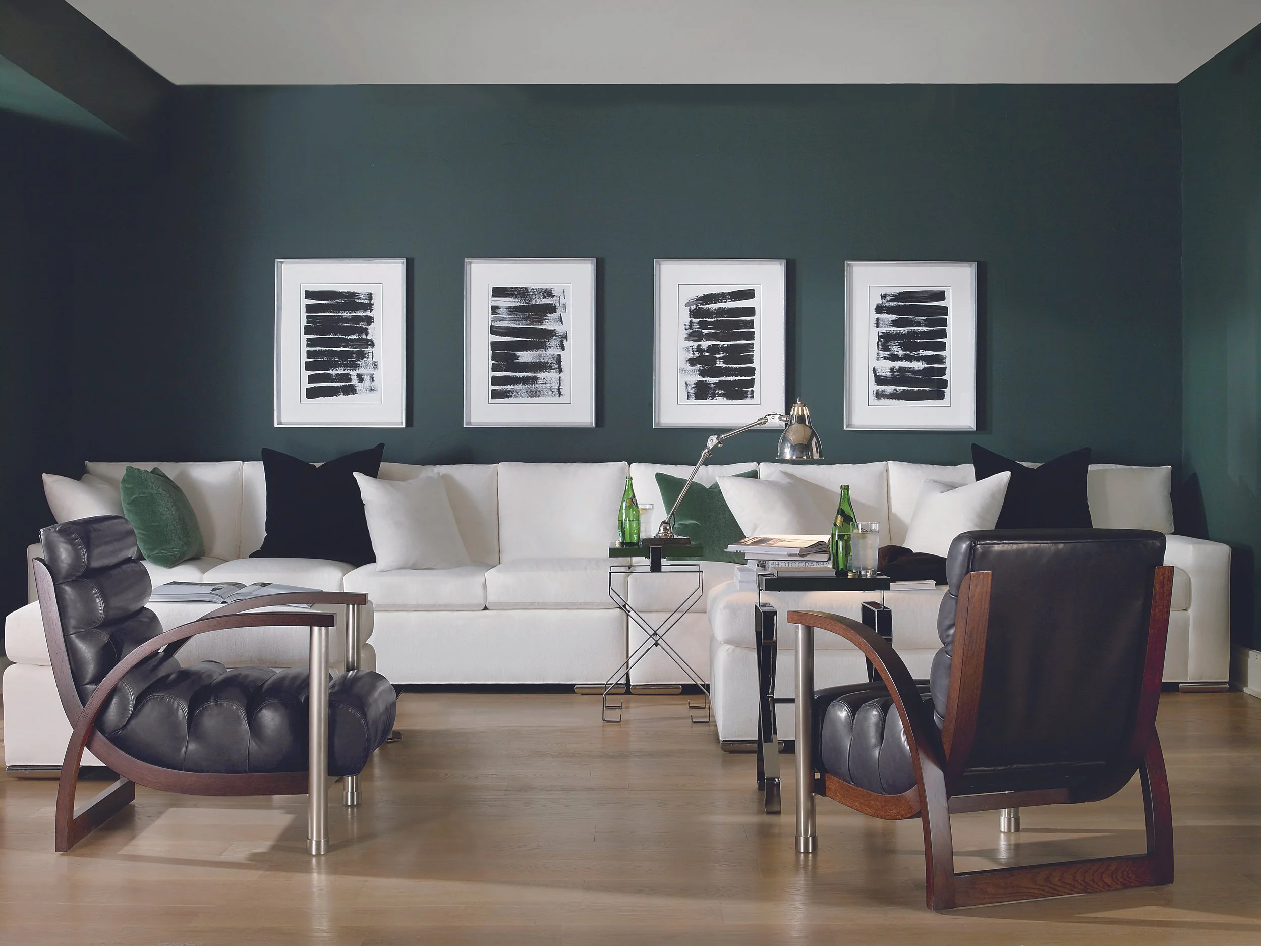 Living room with a white sectional sofa, black and green pillows, dark wall with four black and white abstract framed art pieces, two black leather armchairs, glass side tables with bottles and glasses, a metallic desk lamp, and hardwood flooring.