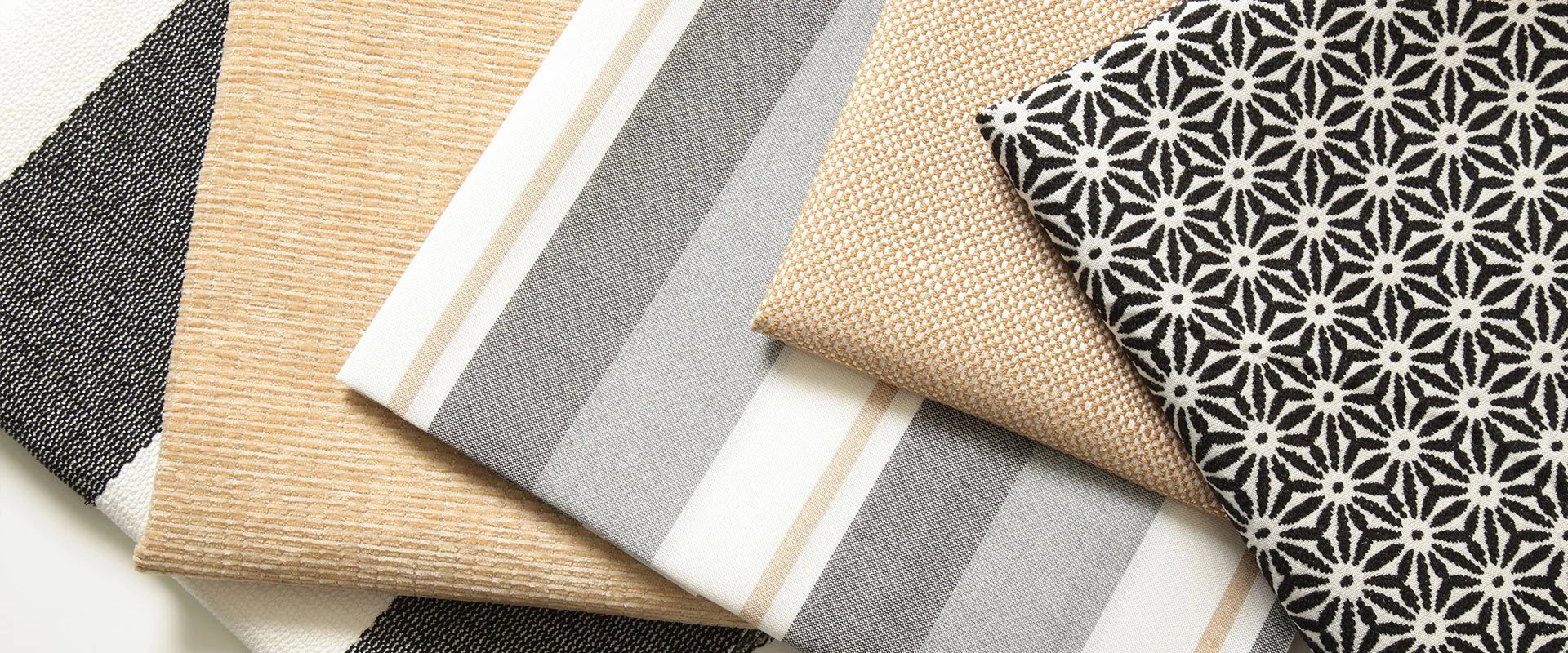 Stacked fabric samples with various patterns and colors, including black and white floral, beige, gray, and striped designs.