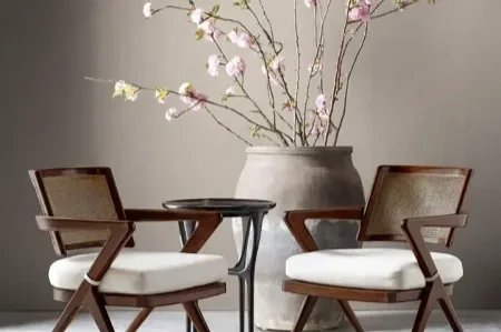 Two mid-century modern chairs with wooden frames and white cushions facing each other with a small round table between them, and a large vase with branches of pink and white flowers behind them.