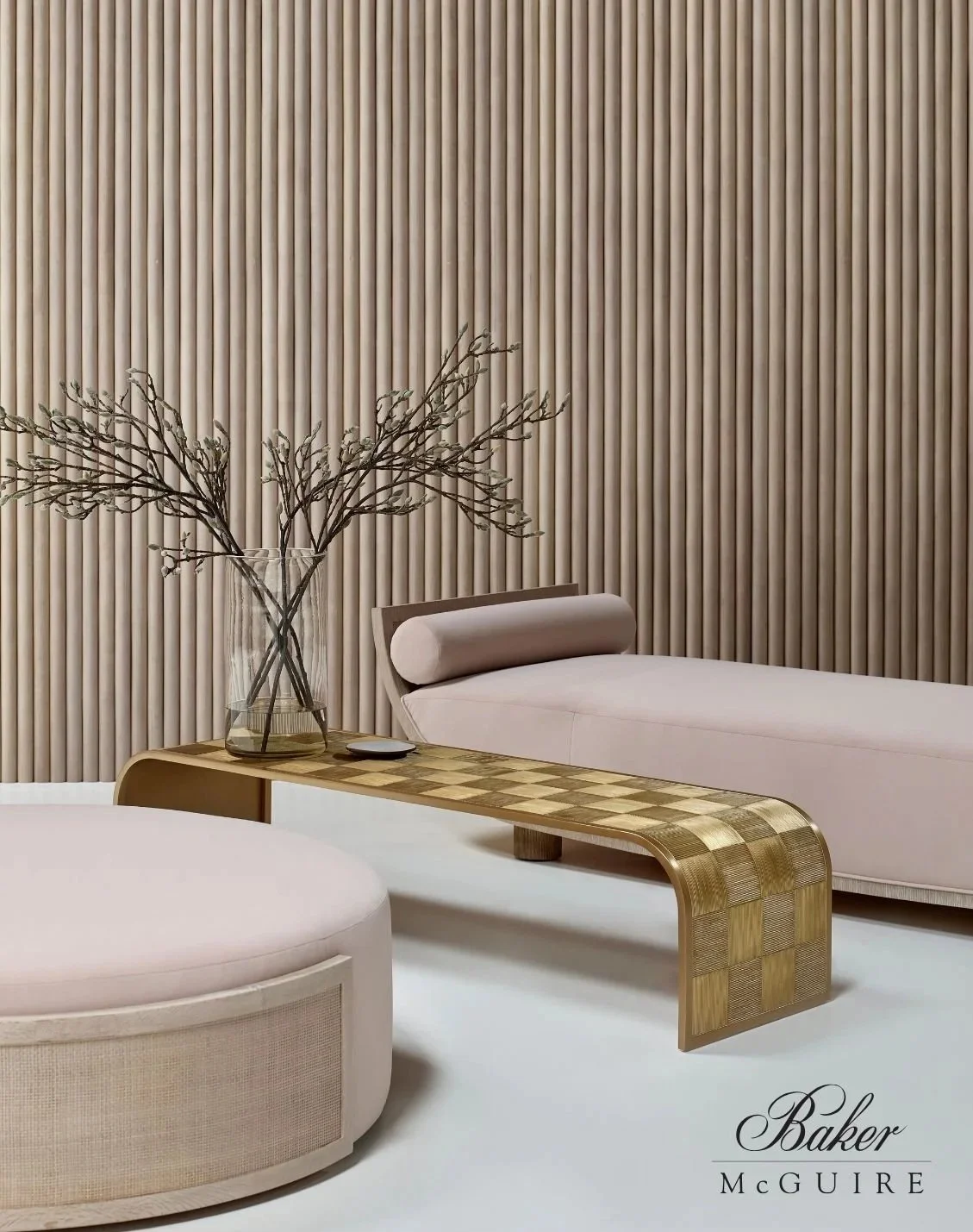 A modern interior room with a beige and cream color palette, featuring a light pink lounge chair with a rolled headrest, a gold-tone bench with a checkered pattern, a glass vase with bare branches, and a wood-paneled wall.