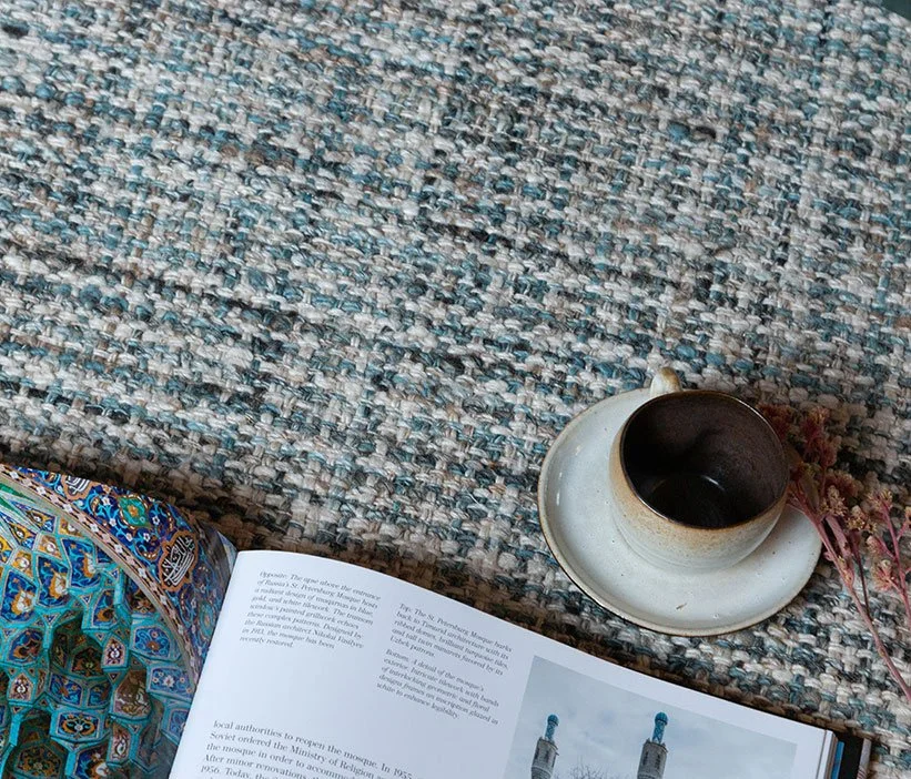 A book open on a textured, multicolored woven rug, with a white ceramic mug on a small saucer and some pinkish-red dried flowers beside it.