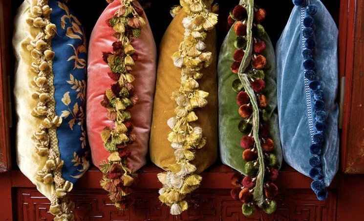 Six colorful decorative pillows with floral and pom-pom trim, arranged on a wooden surface.