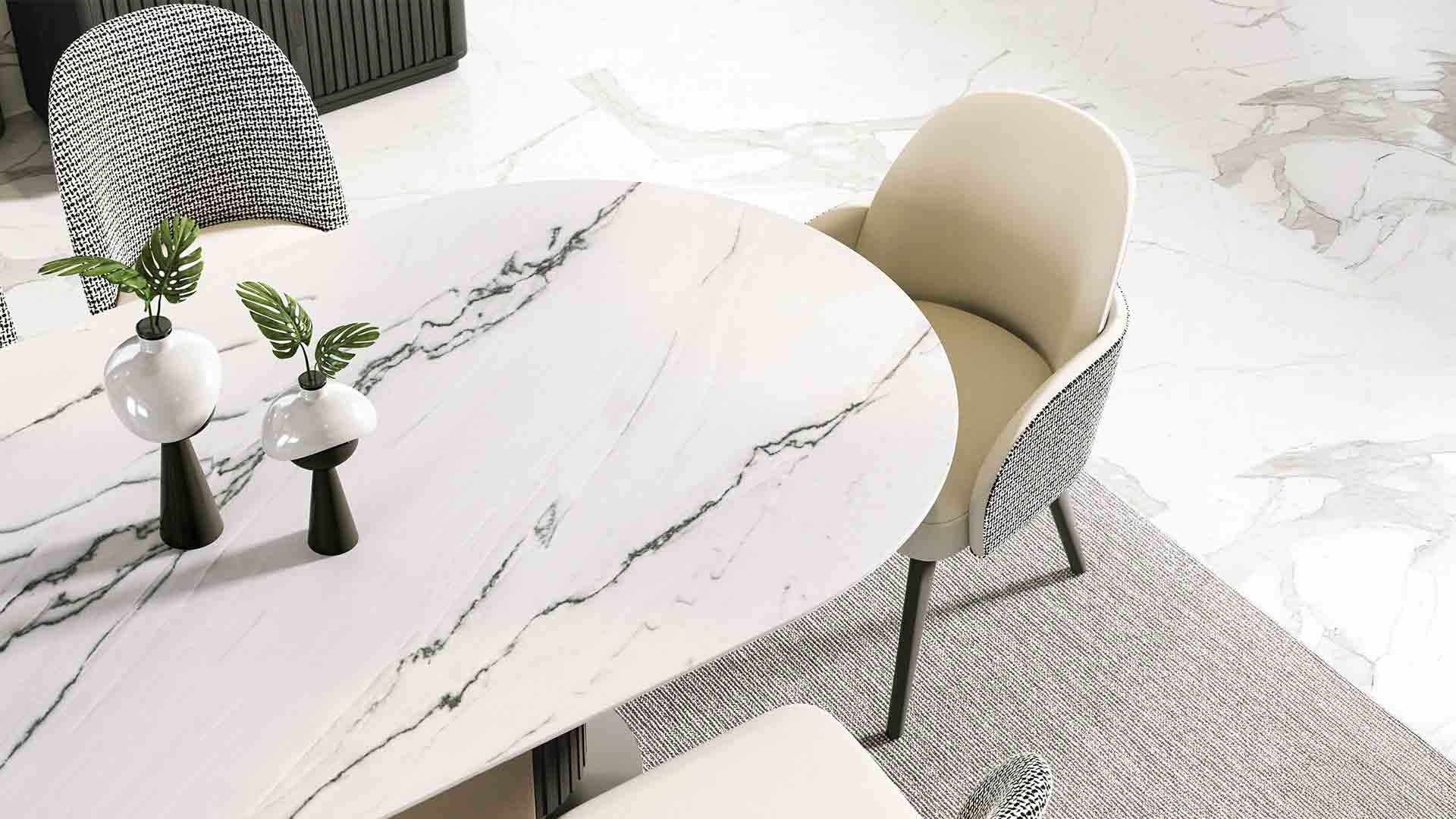 A round white marble dining table with black and white vases and green leaves, surrounded by beige and black patterned chairs, on a marble floor with a textured beige rug underneath.