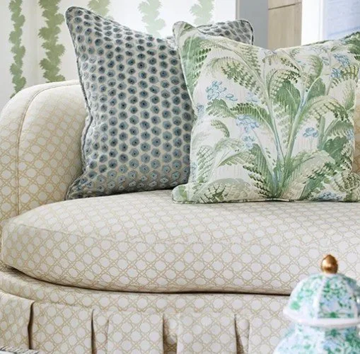 Close-up of a beige sofa with two decorative pillows featuring different patterns and colors, and a small teapot in the bottom right corner.