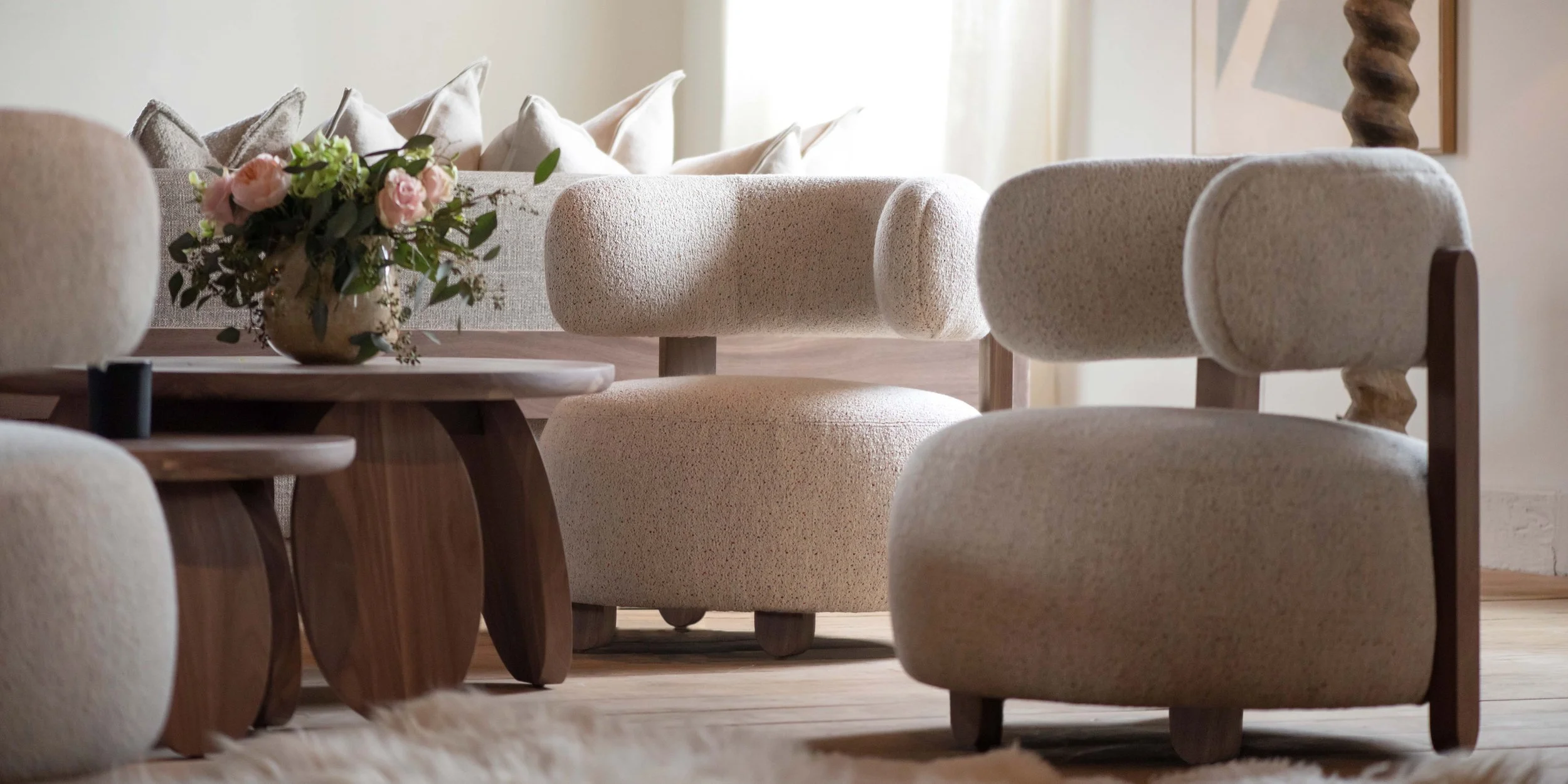 A cozy living room with a beige sofa, a beige armchair, a wooden coffee table, and a vase of pink flowers, with cream-colored pillows on the sofa and natural light coming in from a window.