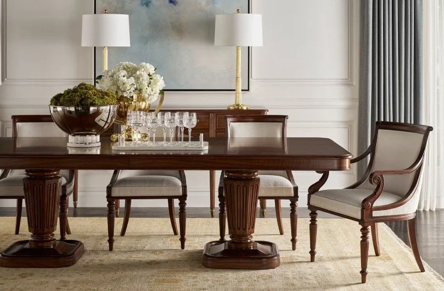 A formal dining room with a dark wood table, six upholstered chairs, and two matching white lamps on a wooden sideboard against a light-colored wall with a framed artwork. A large floral arrangement is on the table, and the room has curtains on the right side.