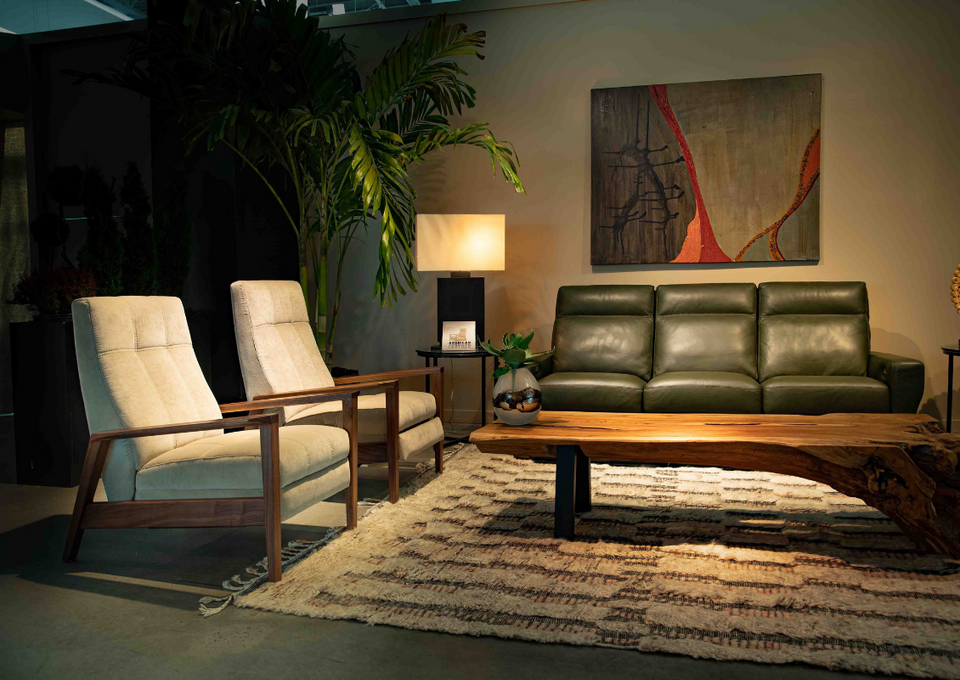Living room with two beige armchairs, a dark green leather sofa, a wooden coffee table, a patterned area rug, a large potted plant, a lamp, and wall art.