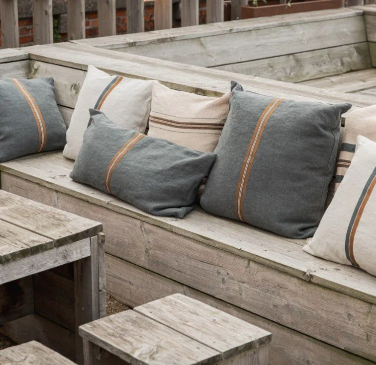 Outdoor wooden bench with multiple decorative pillows in grey, white, and beige with striped patterns.