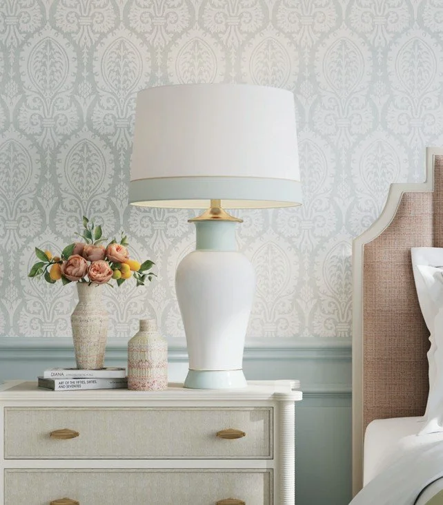 A bedroom nightstand with a white pleated drawer, a large white and light blue ceramic table lamp with a white lampshade, a vase of pink and yellow flowers, a smaller vase, and a stack of books, next to a beige upholstered headboard with a white pillow and bedding against a blue and white damask wallpaper background.