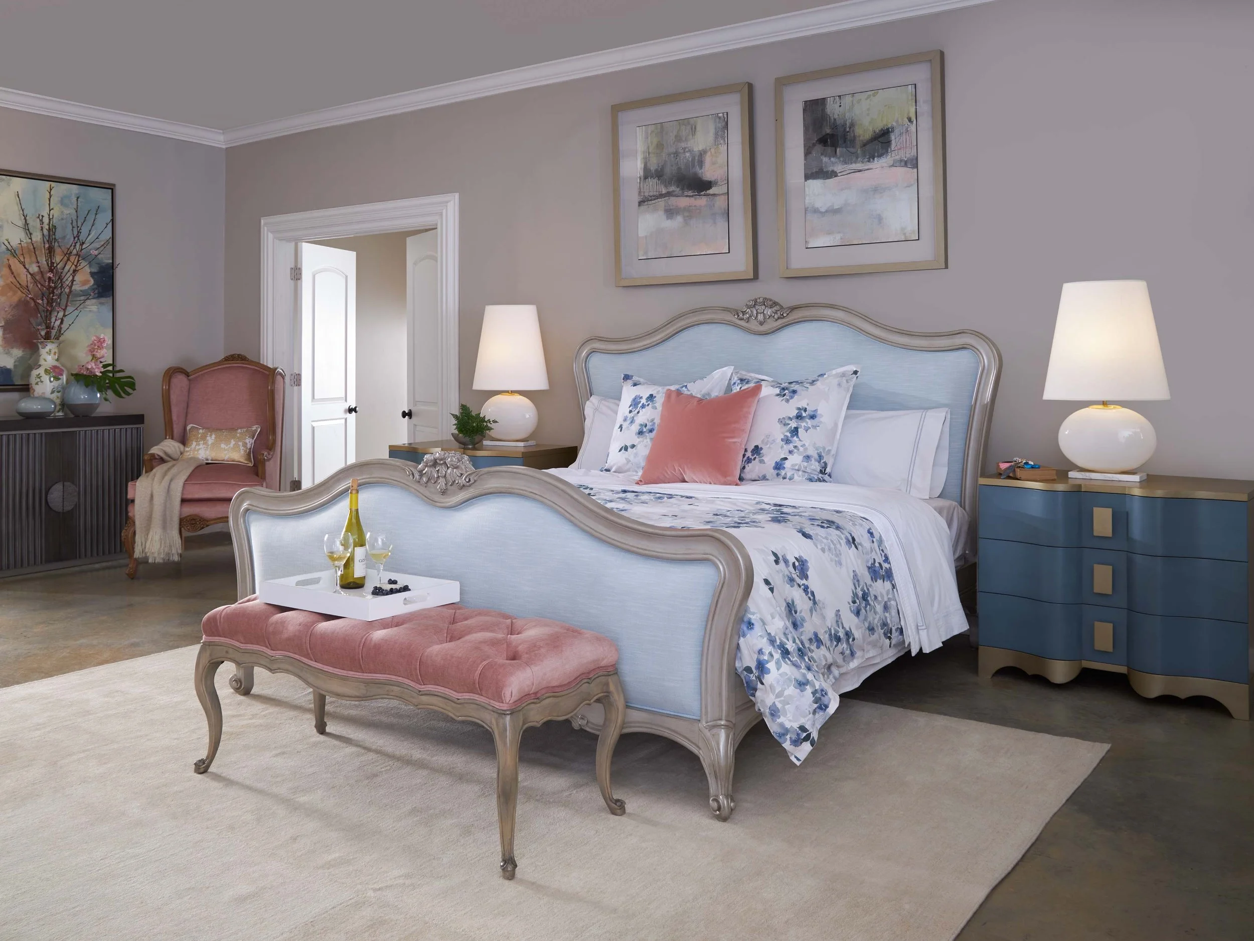 Elegant bedroom with a light blue and beige bed, pink and blue pillows, white and blue floral bedding, two blue nightstands with white lamps, a pink velvet settee with a tray holding wine and grapes, a pink and beige armchair with a beige throw, and wall art with abstract paintings.