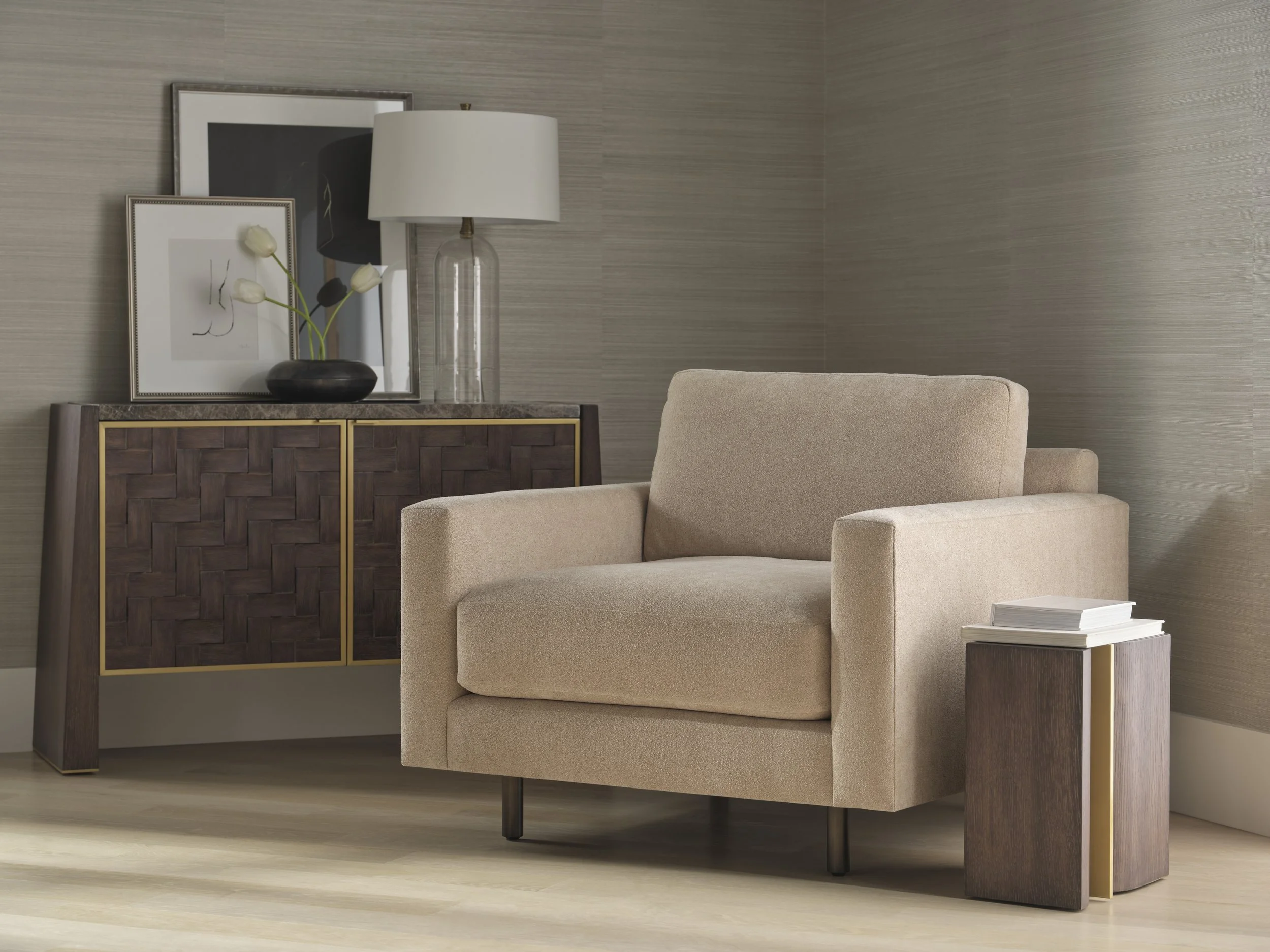 A beige armchair next to a side table with stacked books, and a wooden sideboard with decorative art, a table lamp, and a vase with white tulips, in a modern interior space.