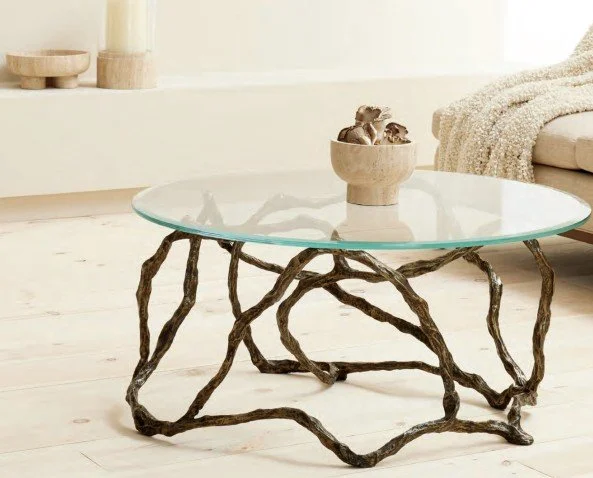 Round glass coffee table with a twisted, branch-like wooden base in a bright room, with a beige bowl filled with decorative items on top.