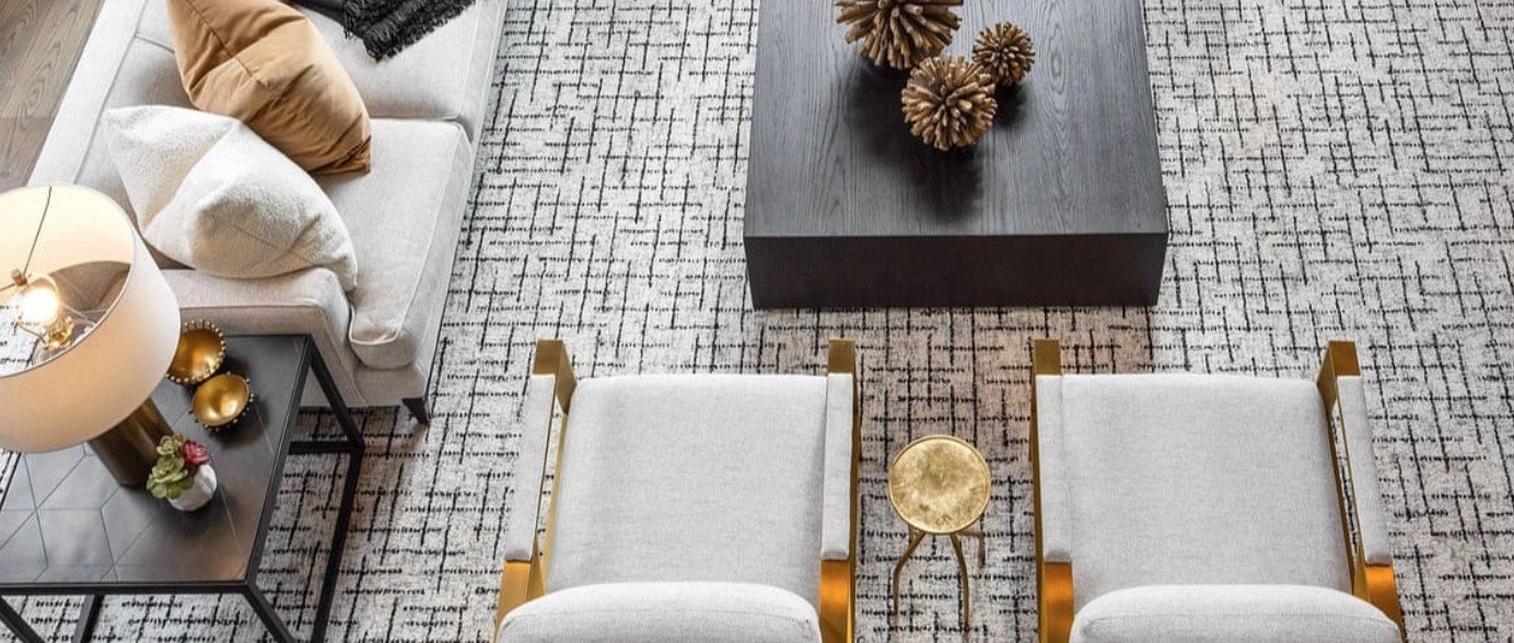 Overhead view of a modern living room with a white sofa, black side table with gold decorative items, a patterned rug, two white chairs with gold accents, a black coffee table with decorative objects, a round side table, and a lamp.
