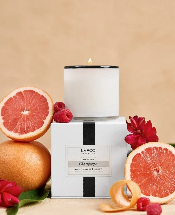 A white candle with a lit flame, placed on top of a box, surrounded by grapefruit halves, raspberries, red flowers, and a grapefruit with a peel.