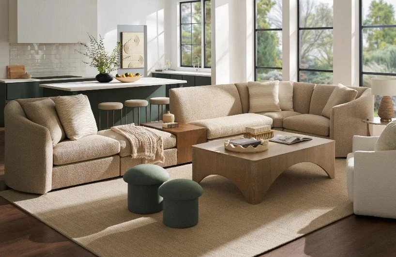 A modern living room with beige sofas, a wooden coffee table, green ottomans, and large windows letting in natural light, overlooking trees outside.