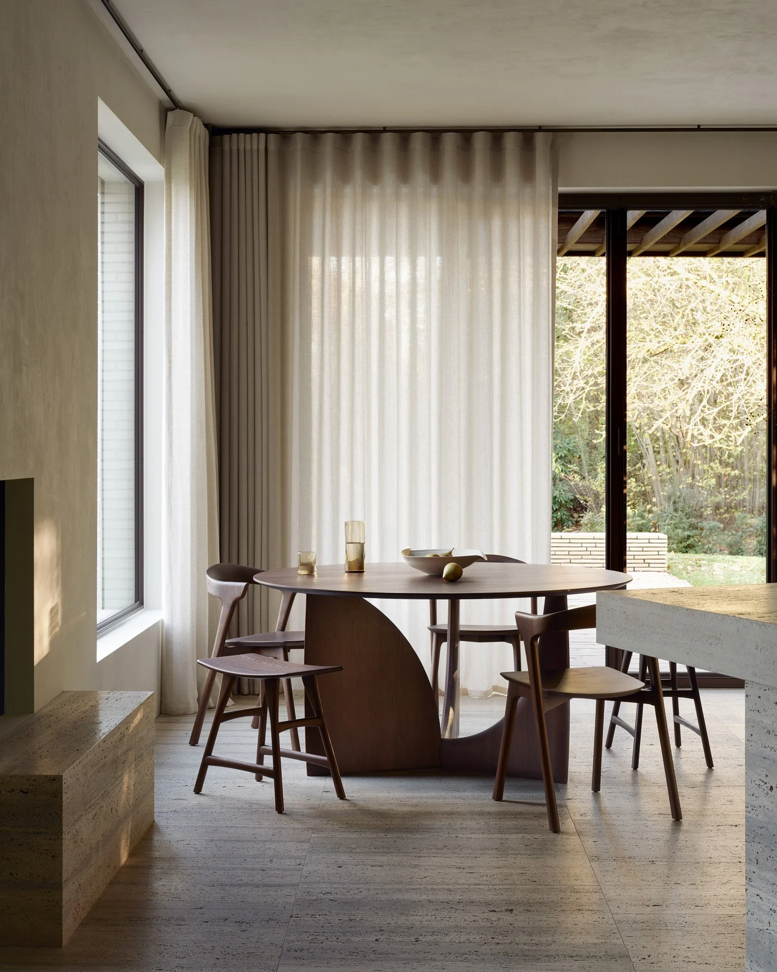Modern dining area with round wooden table, four chairs, large window with sheer curtains, and sliding glass door showing outdoor garden.