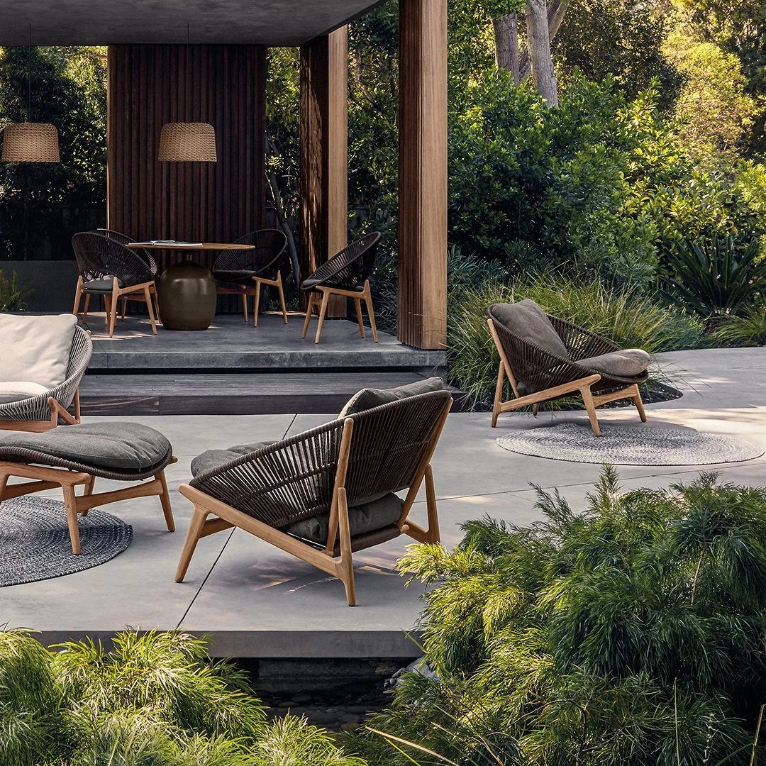 Outdoor patio with modern furniture, including cushioned chairs and a small table on a concrete surface, surrounded by lush green bushes and trees, with a covered seating area in the background.