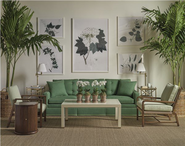 Living room with green sofa, framed botanical artwork on light green wall, large potted palm trees on either side, wooden chairs, white coffee table with potted plants, side tables with lamps, and woven baskets.