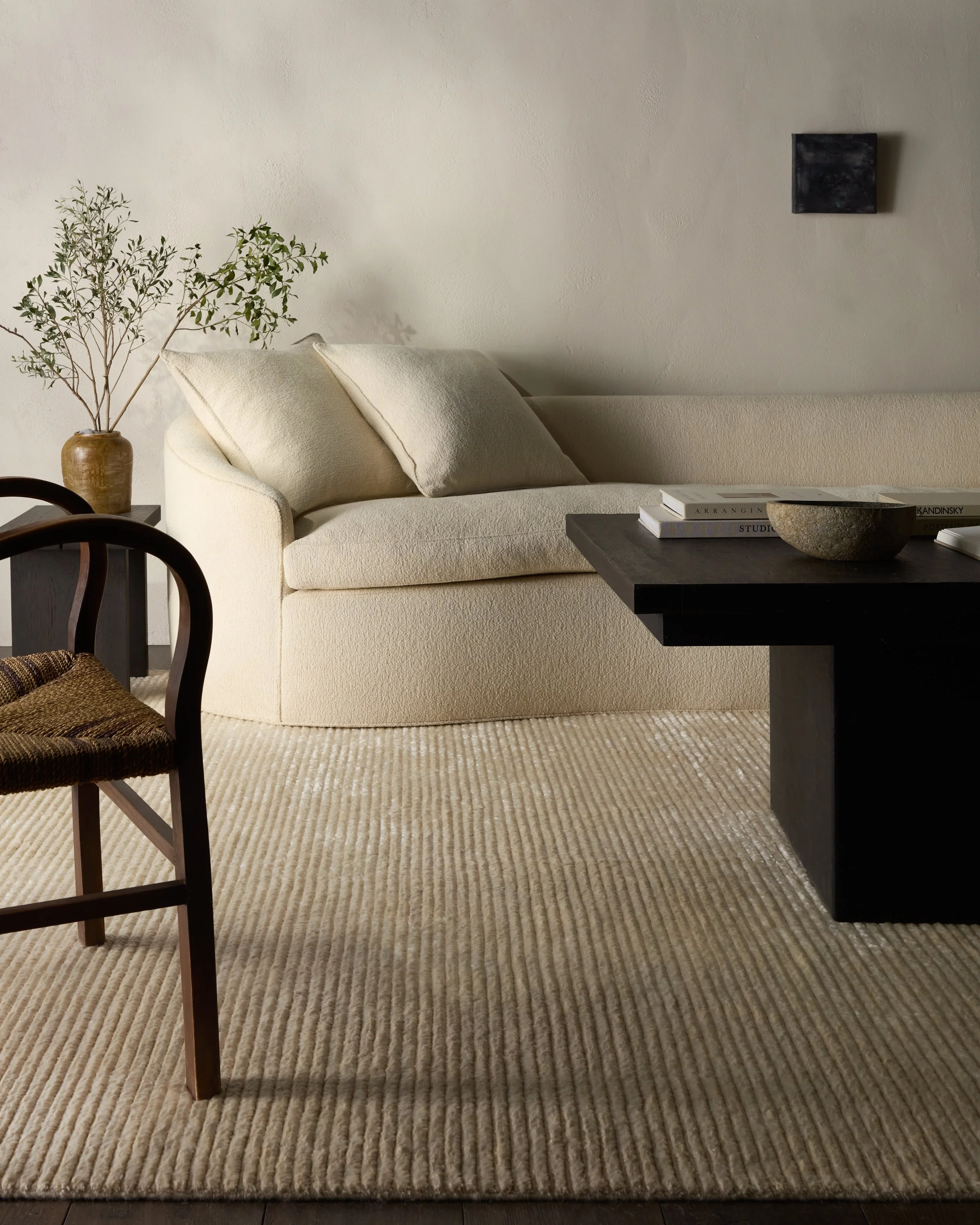 Living room with a cream-colored sofa, a dark coffee table with books and a bowl, a woven chair, a plant in a pot, and minimalist wall art on a textured wall and beige carpet.