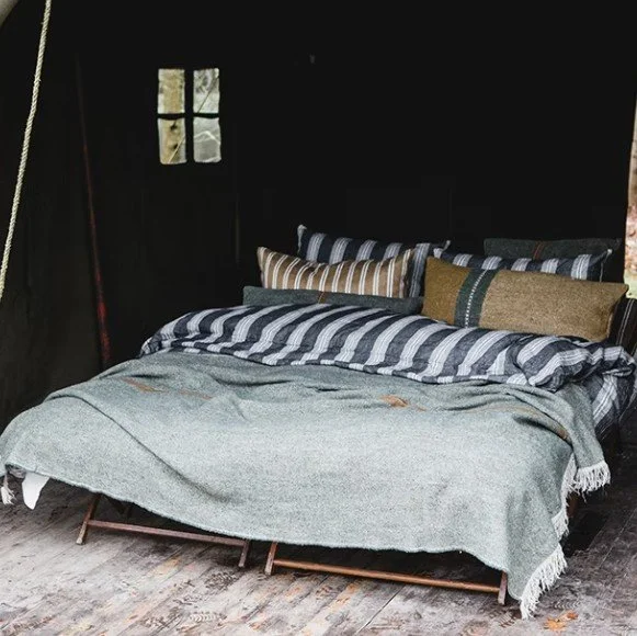 A bed with multiple striped and solid-colored pillows inside a rustic, dark cabin with a small window.