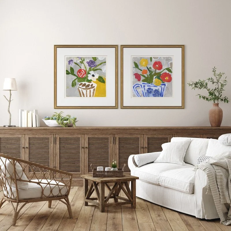 Living room with two colorful floral paintings above a wooden cabinet, white sofa, woven armchair, small side table, and houseplants in a neutral decor.