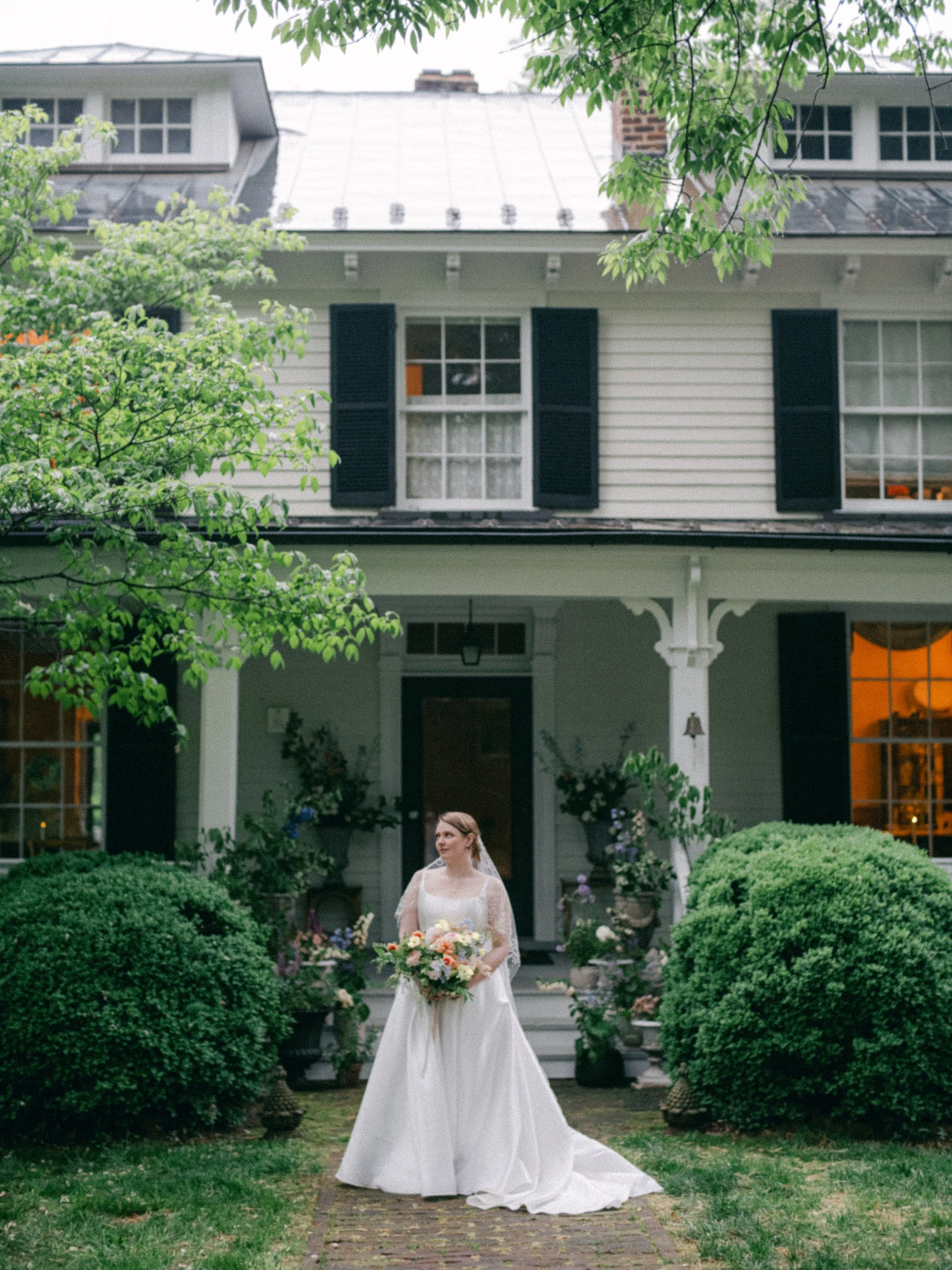 A bride in a white wedding gown holding a bouquet, standing outdoors in front of a house with black shutters, greenery, and flowering bushes.