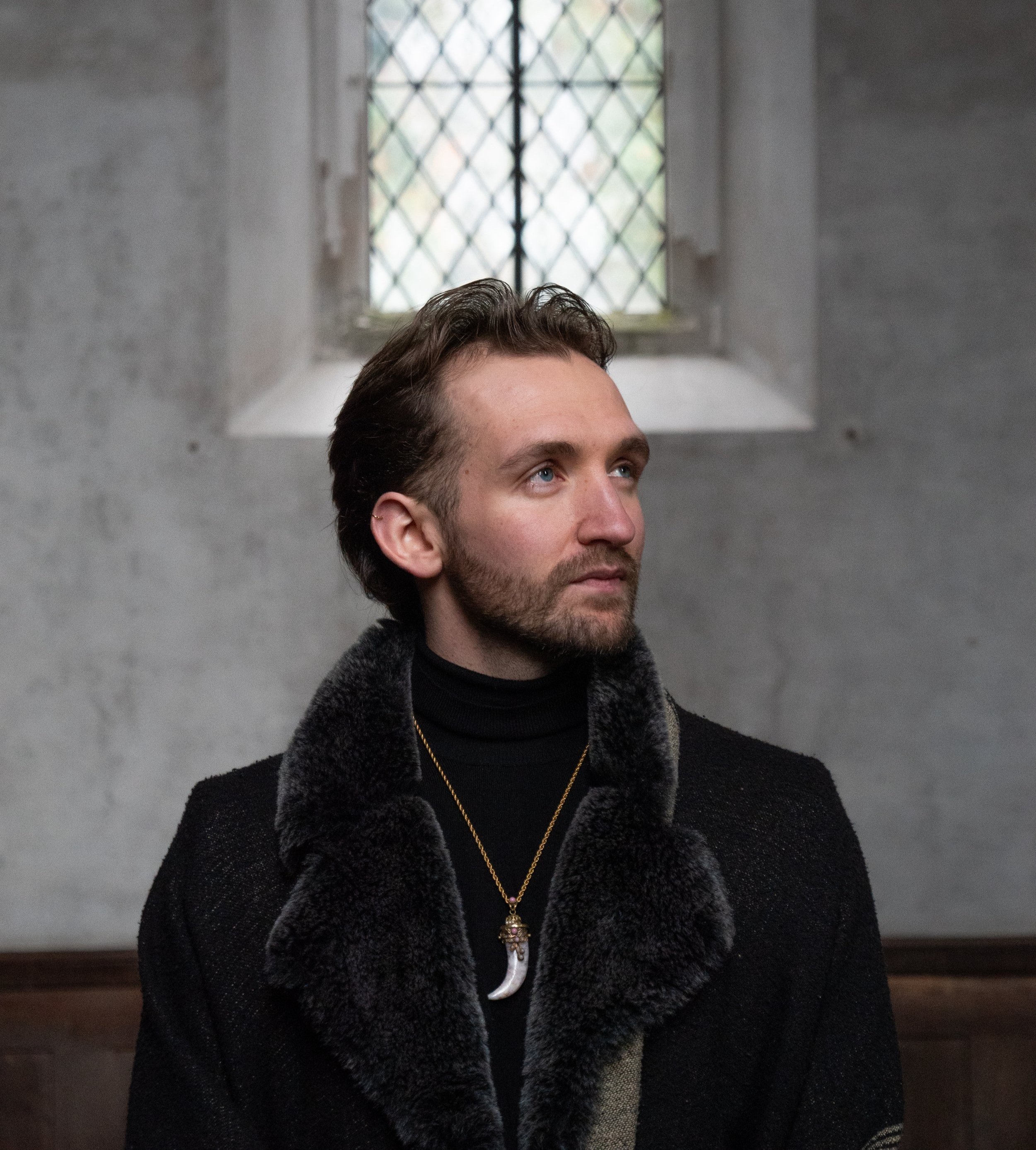 A man with dark hair and a beard wearing a black fur-collared coat and a necklace with a tooth pendant sits inside near a window with diamond-shaped panes.