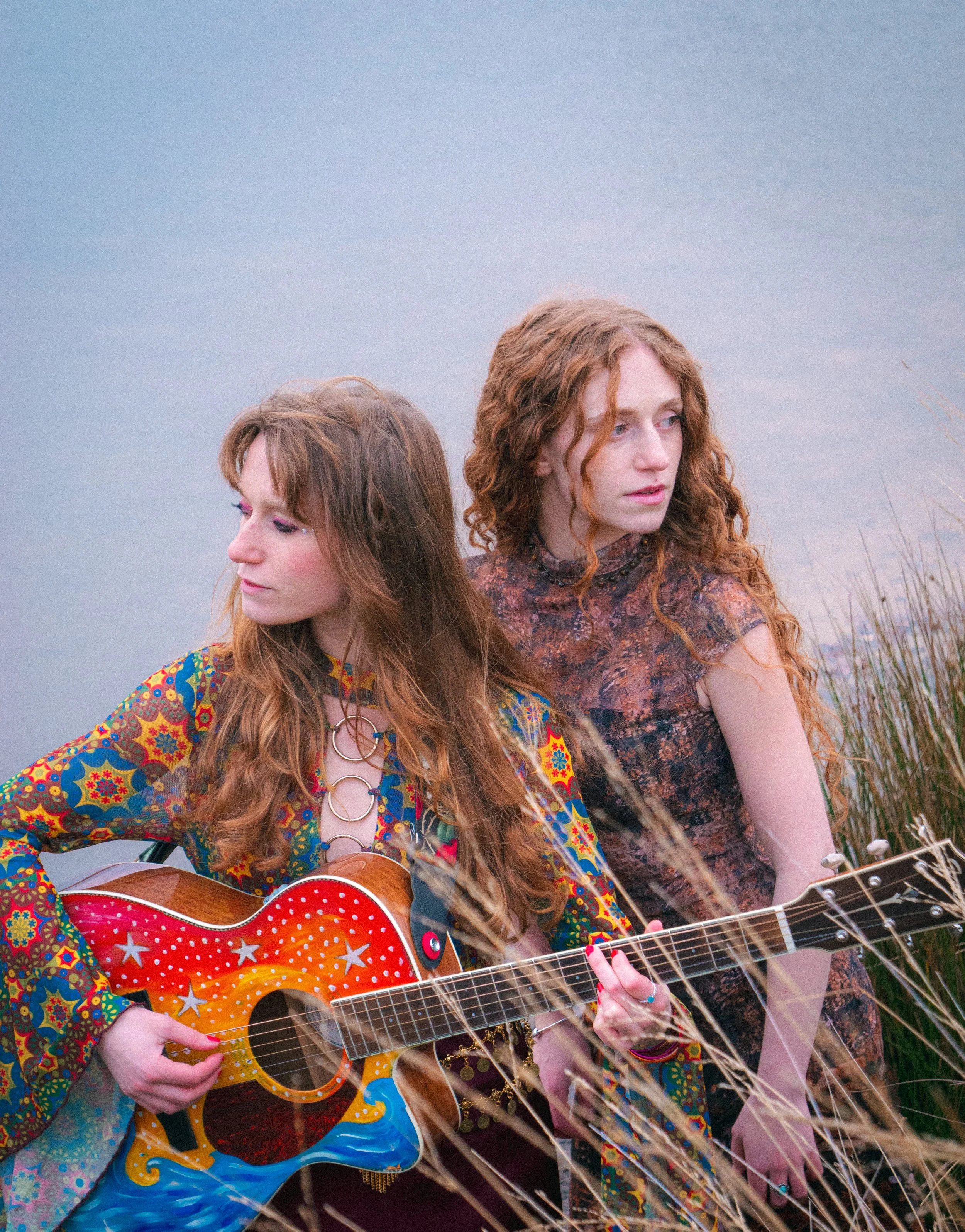 Two women with red hair sitting outdoors in a field of tall grass; one is playing a colorful, star-decorated acoustic guitar, the other is looking away toward the right side of the frame.