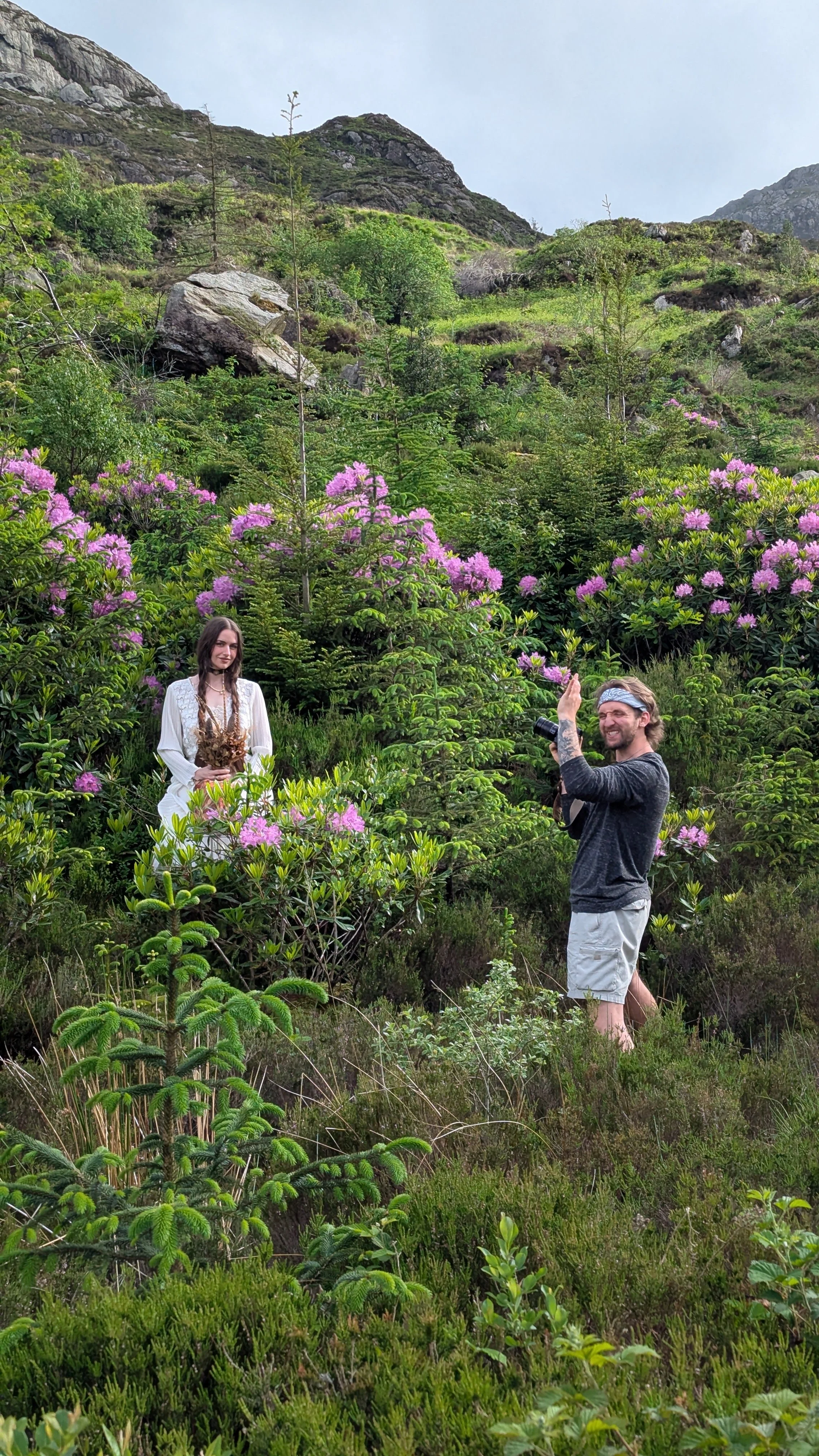 A woman and a man stand amidst green plants and blooming pink flowers on a mountainous hillside. The woman is holding a bunch of flowers, and the man is smiling, holding a camera, and waving.