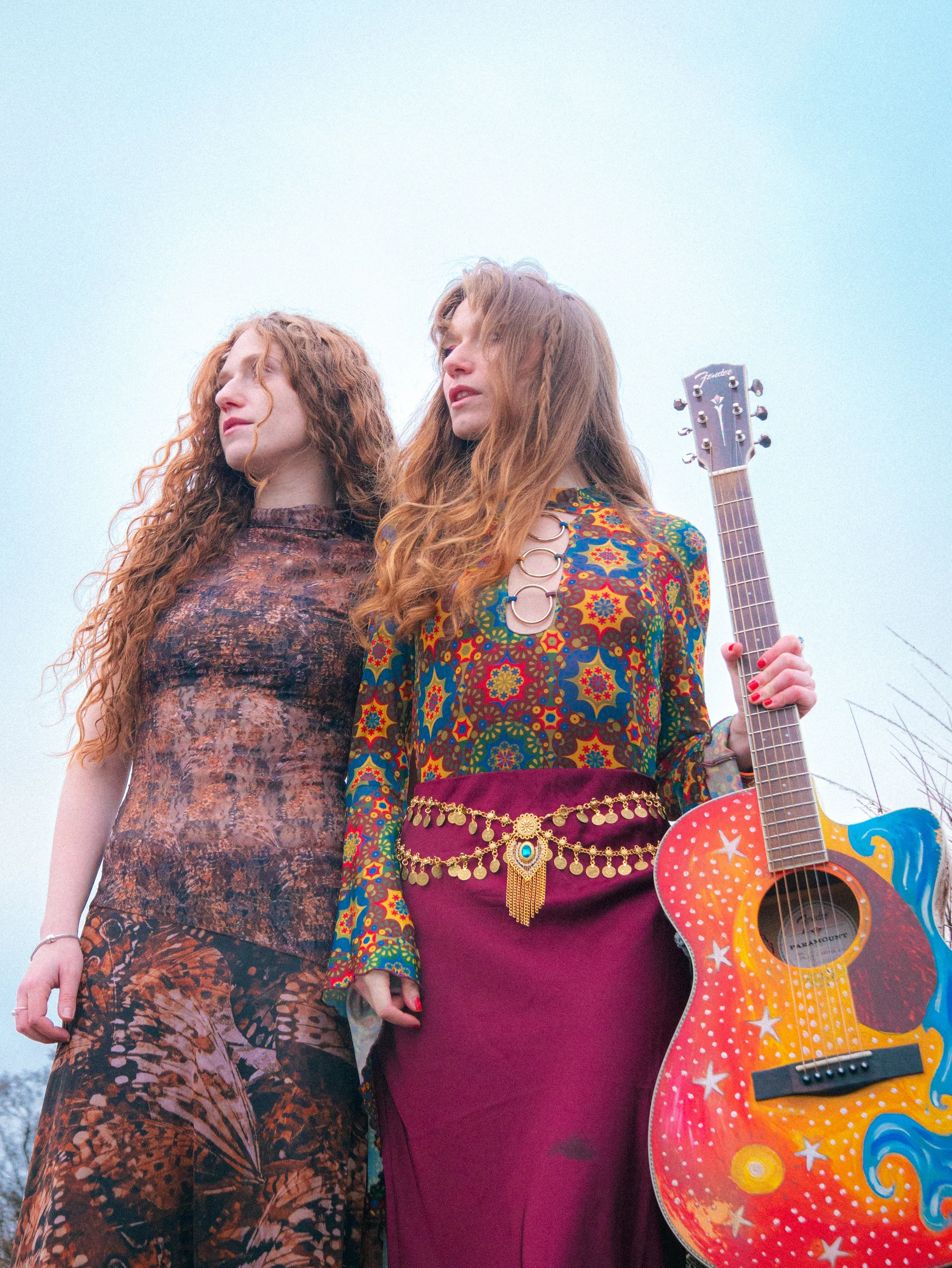Two women with long, red, curly hair standing outdoors against a light blue sky. One woman wears a patterned dress, and the other a colorful, patterned blouse with a purple skirt, holding a decorated red, orange, and yellow acoustic guitar with stars