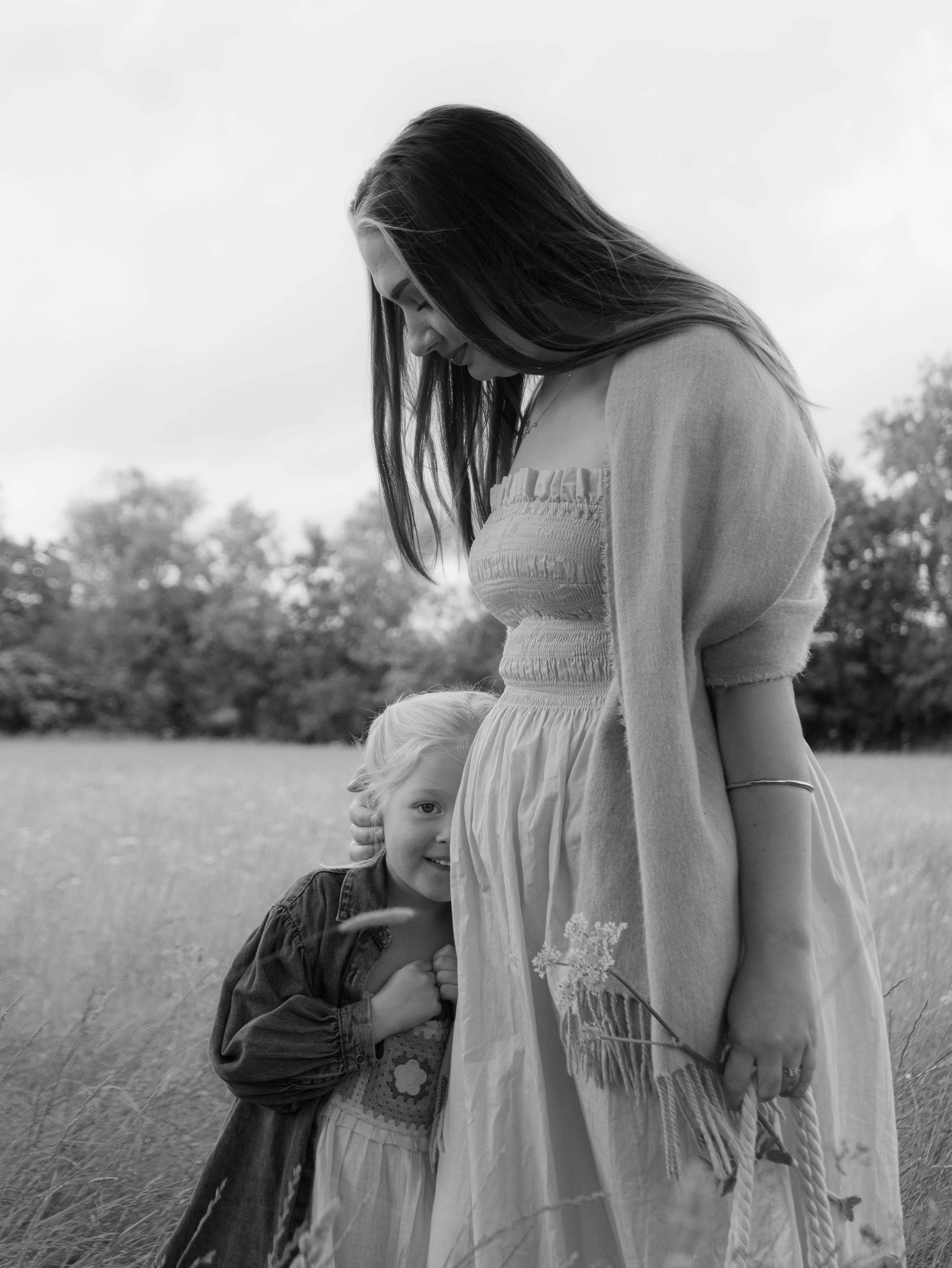 A black and white photo of a woman standing outdoors in a grassy field with a young girl hugging her from the side and looking at the camera. The woman is smiling gently while looking down, and the girl is smiling with her hand on the woman's dress, holding a small flower.