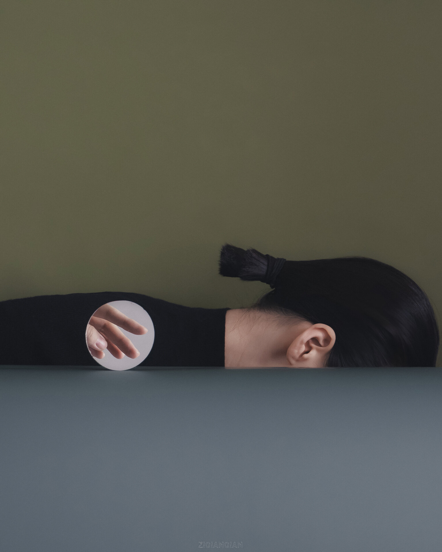 An art photograph that depicts a woman lying face-down, but it appears as though the front half of her body is in the floor, you can only see the back half. There is a small circular mirror beside her with a hand reflected in it.