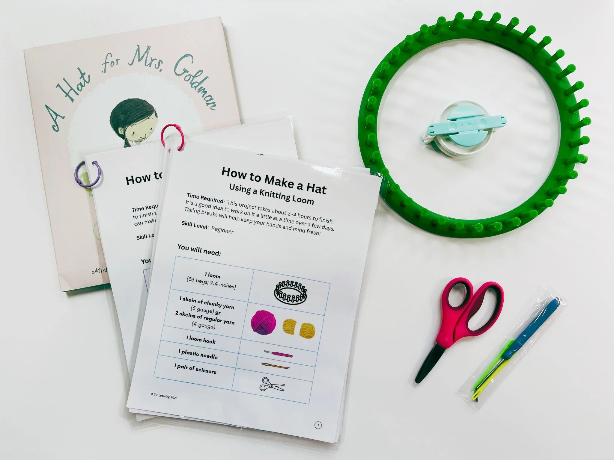 A knitting loom, instruction sheets for making a hat using a knitting loom, scissors, a plastic needle, colorful yarn, and a pair of pink scissors on a white surface.