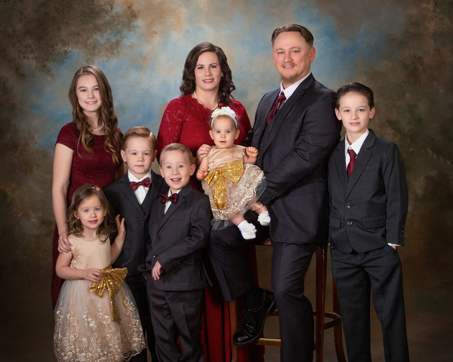 Fine art studio portrait of a large family of eight in coordinating burgundy and black formal attire — parents with six children including a baby