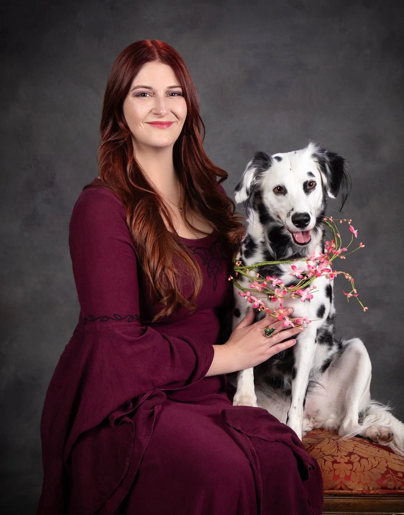 Fine art portrait of a woman with auburn hair in a burgundy dress seated beside her Dalmatian dog holding pink flowers