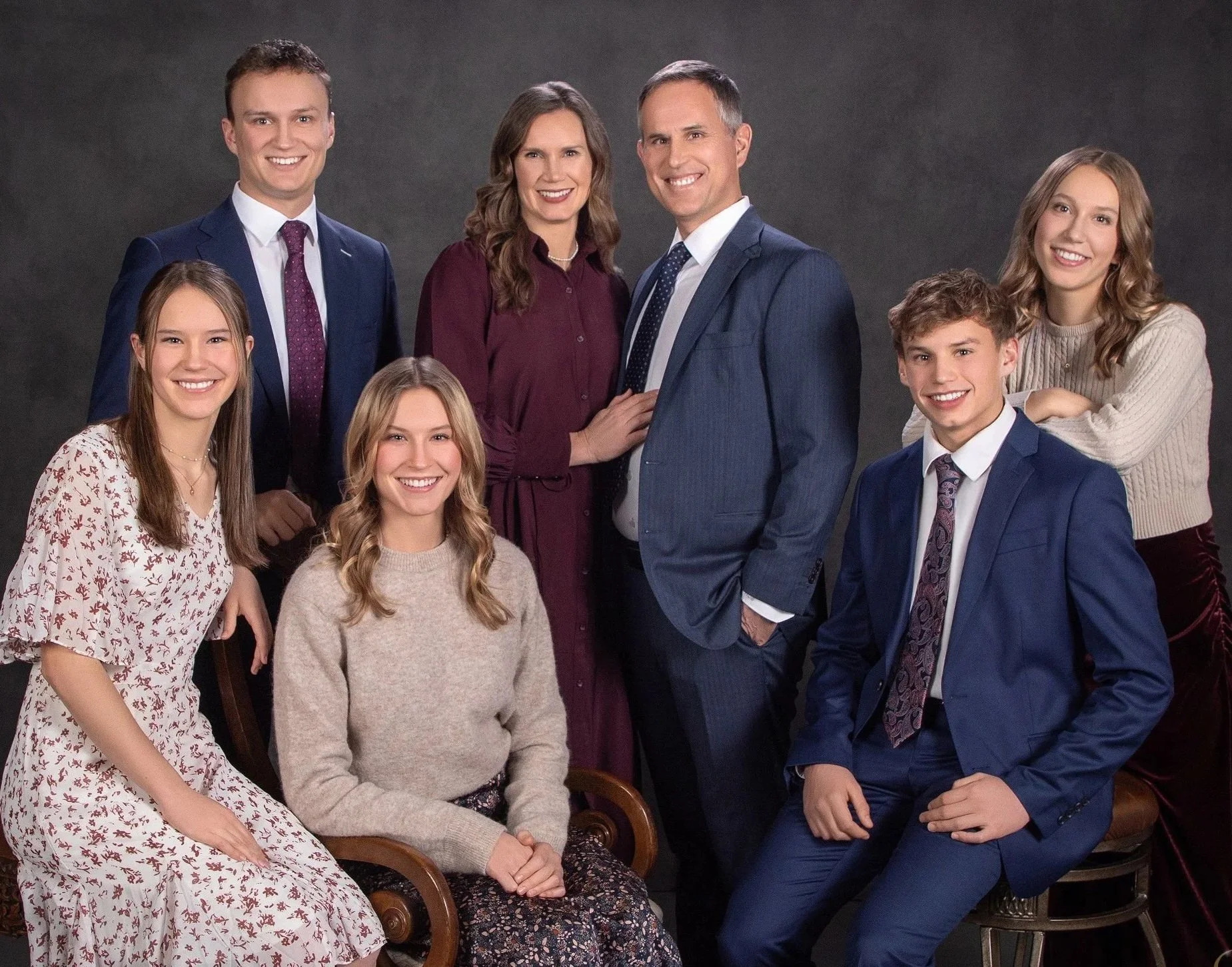 Fine art studio portrait of a family of seven in coordinating navy, burgundy, and cream formal attire against a light painted backdrop