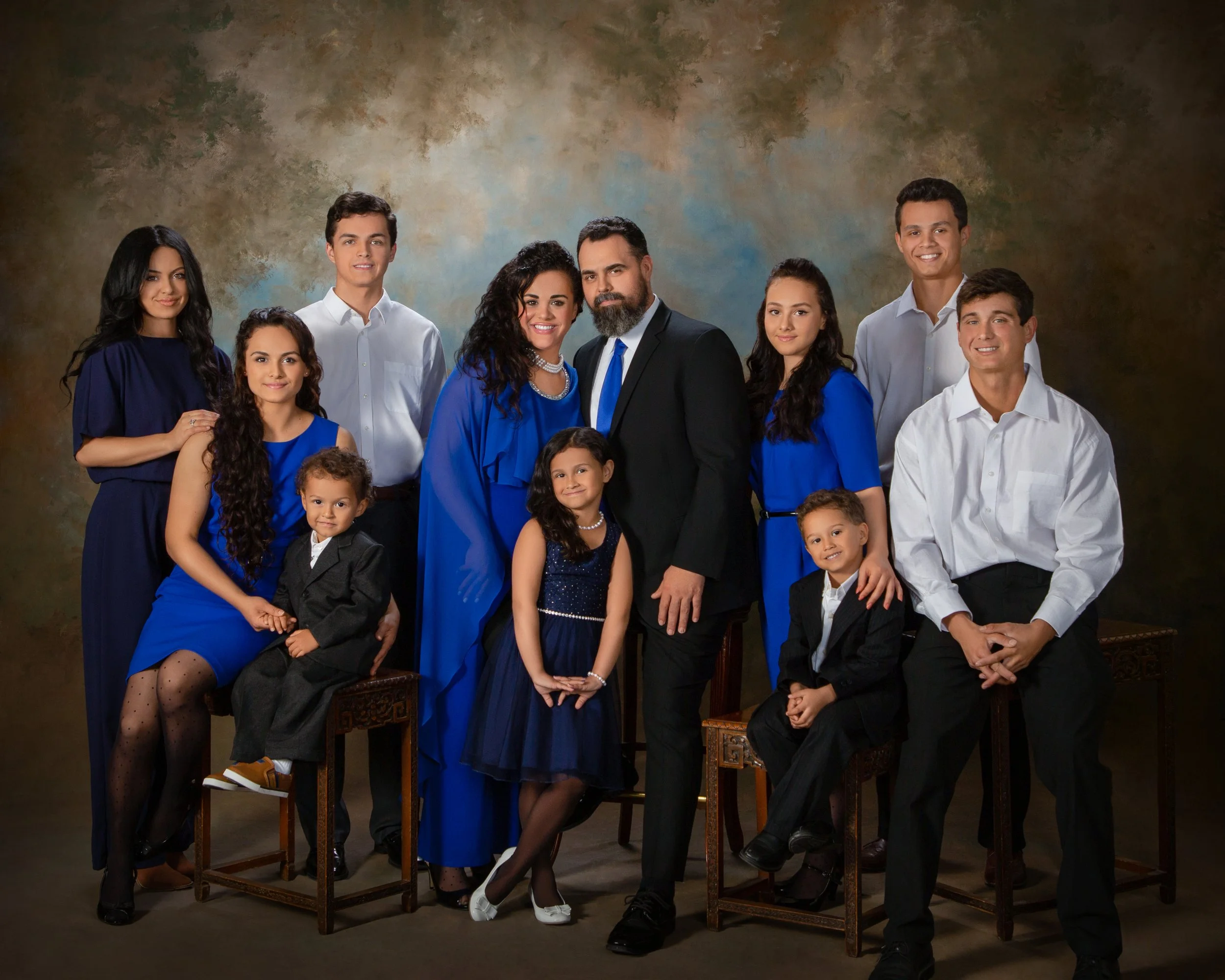 Fine art studio portrait of a large multi-generational family in coordinating royal blue and black formal attire against a painterly backdrop