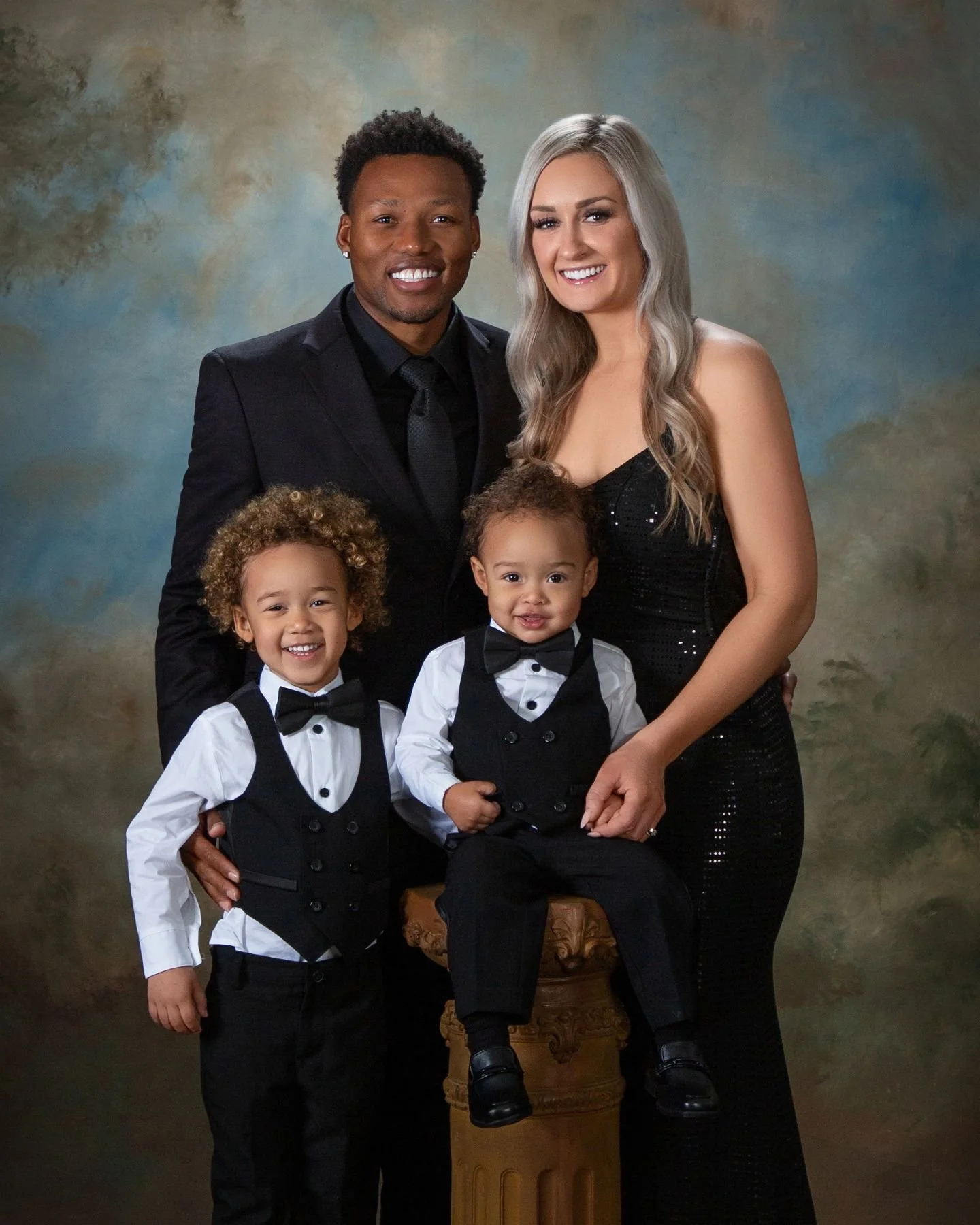 Fine art studio portrait of a family of four in coordinating black formal attire — parents with two young sons in tuxedo vests and bow ties against a painterly backdrop