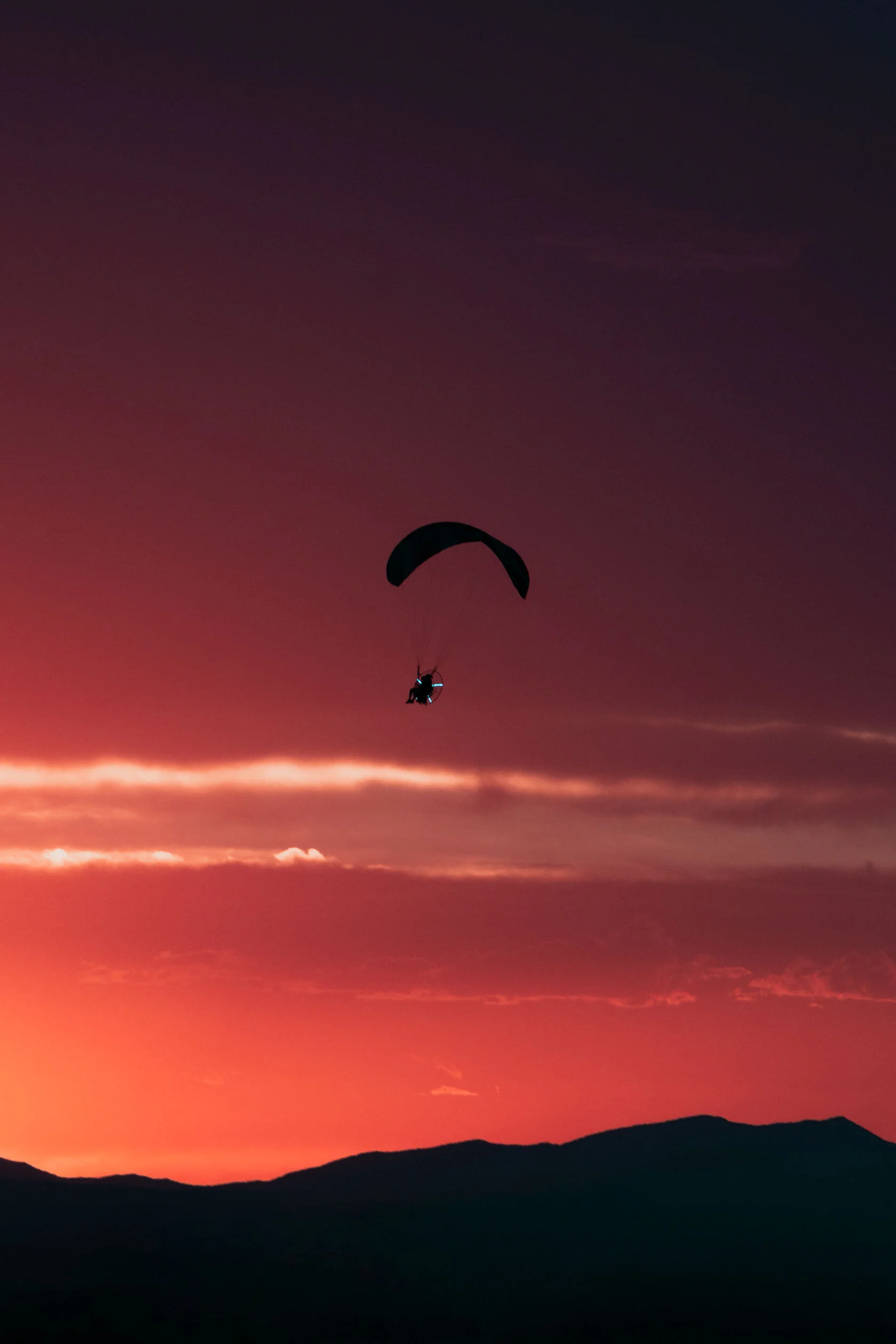 Dark silhouette of guy wearing a motor with lights on it attached to paraglider flies over black, jagged mountains into colorful, dark blue to red orange gradient sunset. In Pocatello, Idaho.