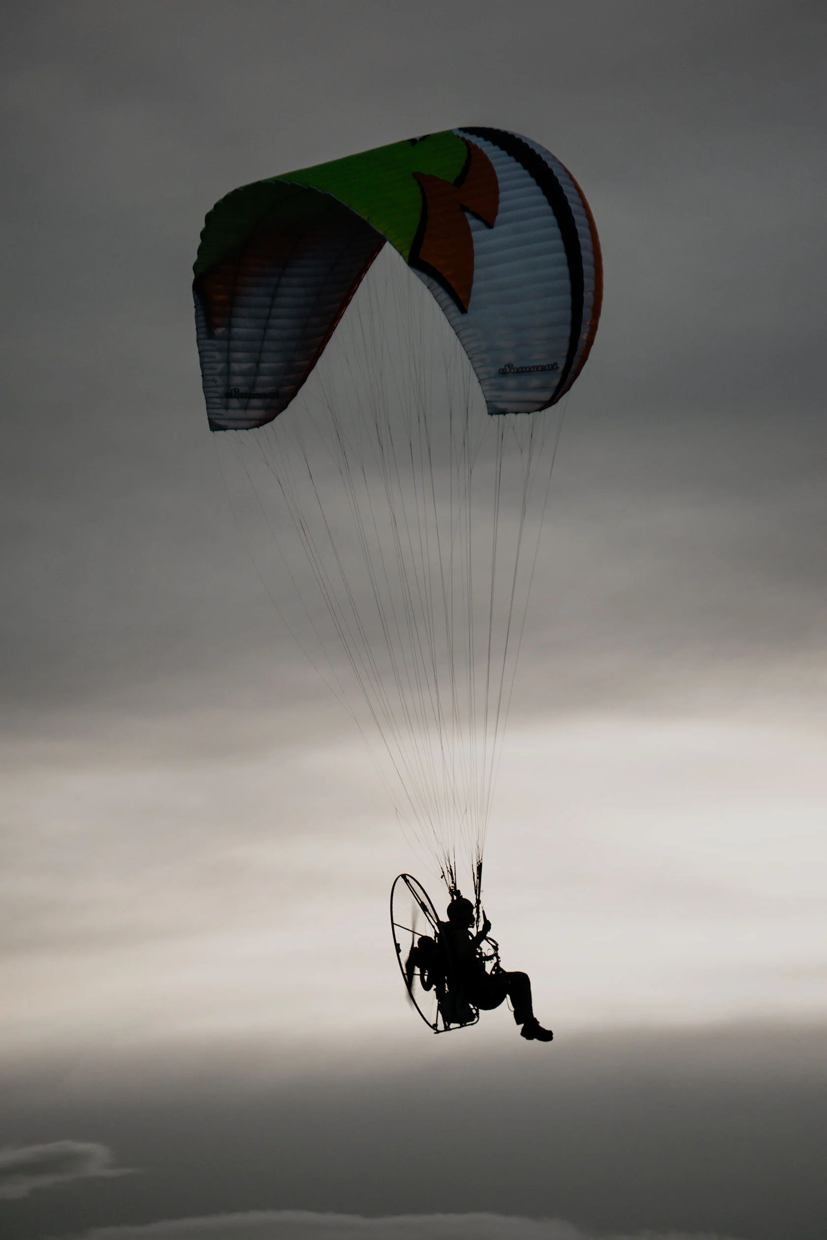 Silhouette of pilot in profile wearing motor attached to green, white, and orange paraglider wing against light white and grey clouds. In Island Park, Idaho.