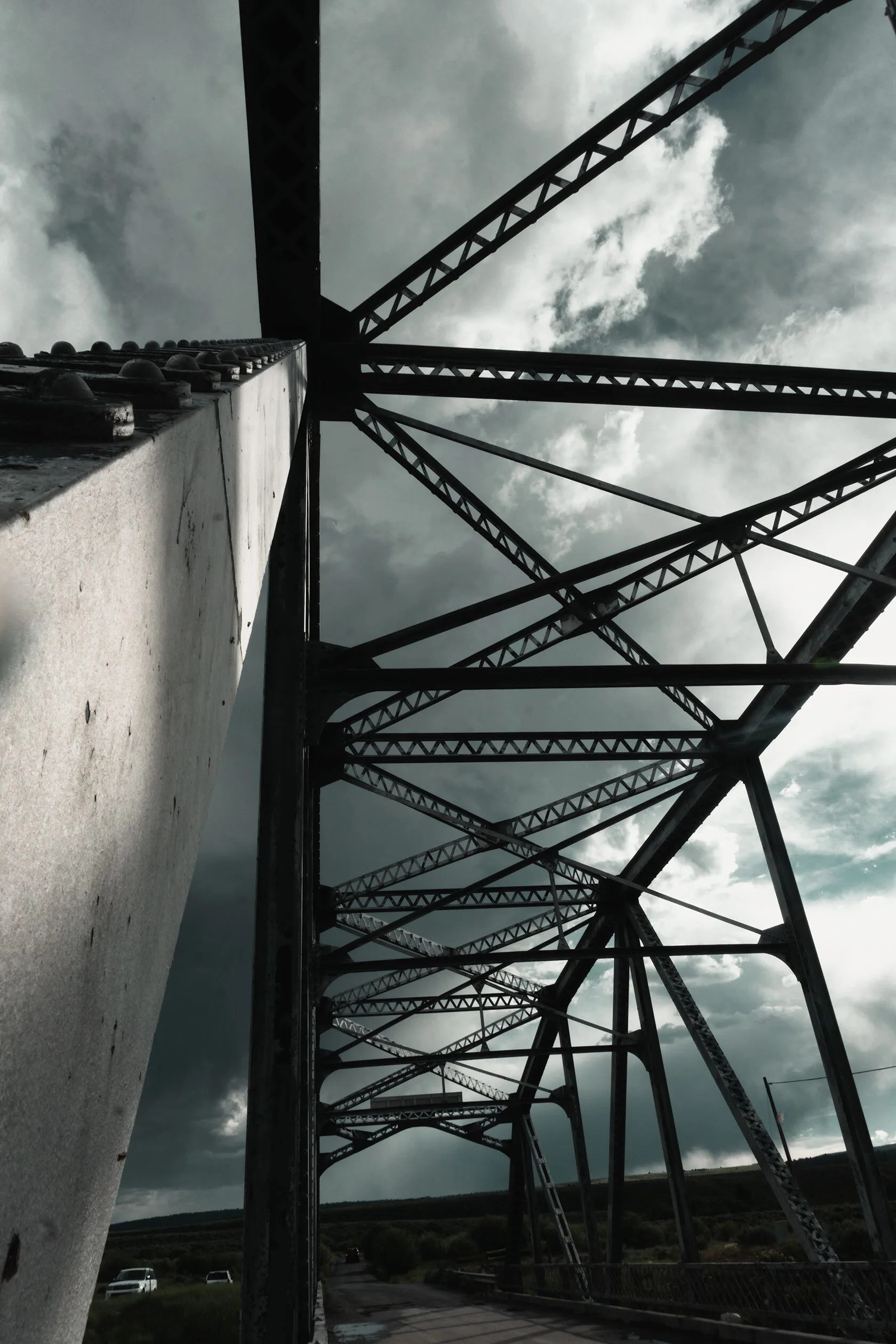 Lopsided view of a beam from a metal suspension bridge leading into a silhouette of the top of the bridge against fluffy storm clouds somewhere in Montana.