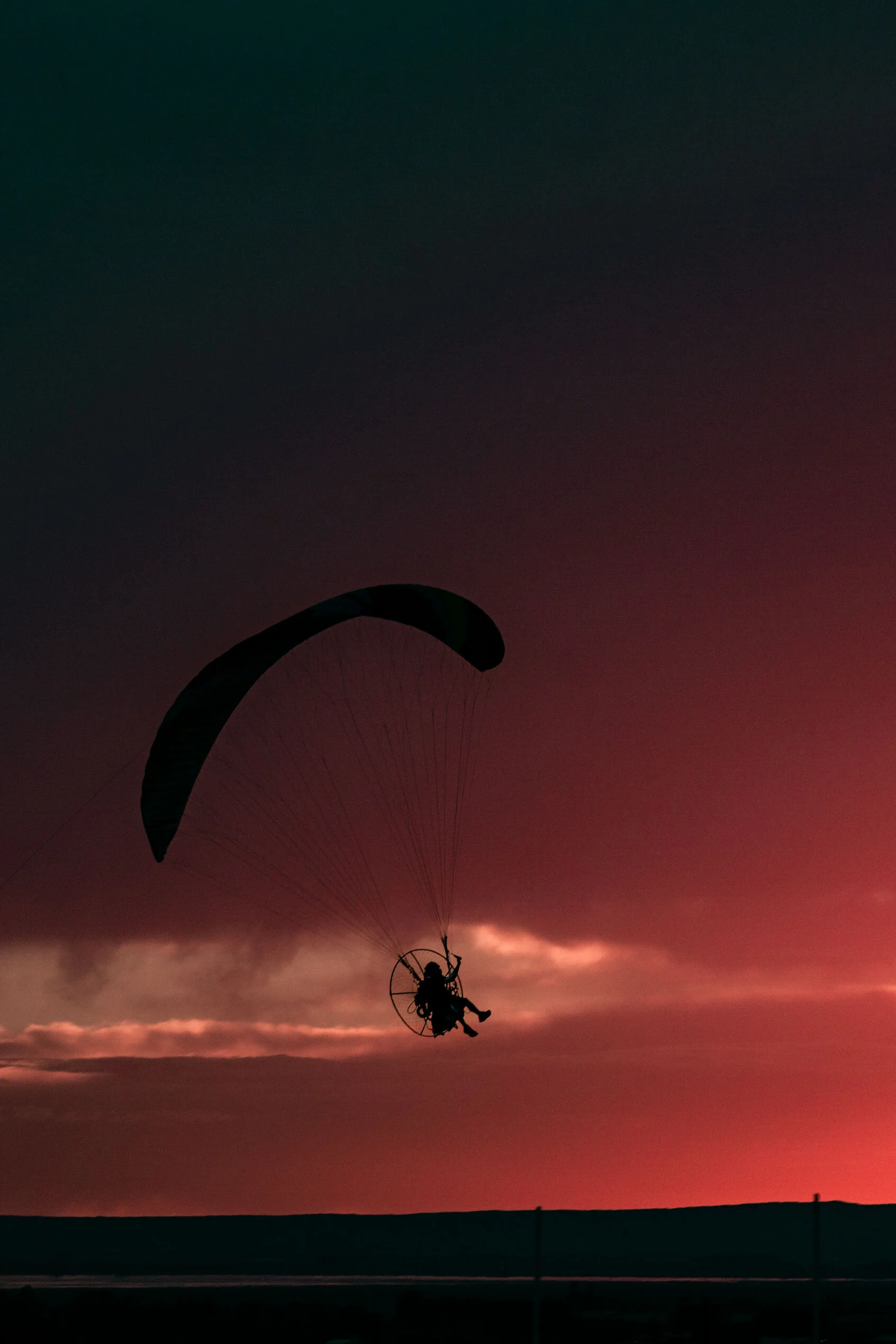 Dark silhouette of guy wearing a motor attached to paraglider flies over black, rolling hills into bright red sunset and wispy colorful clouds. In Pocatello, Idaho.