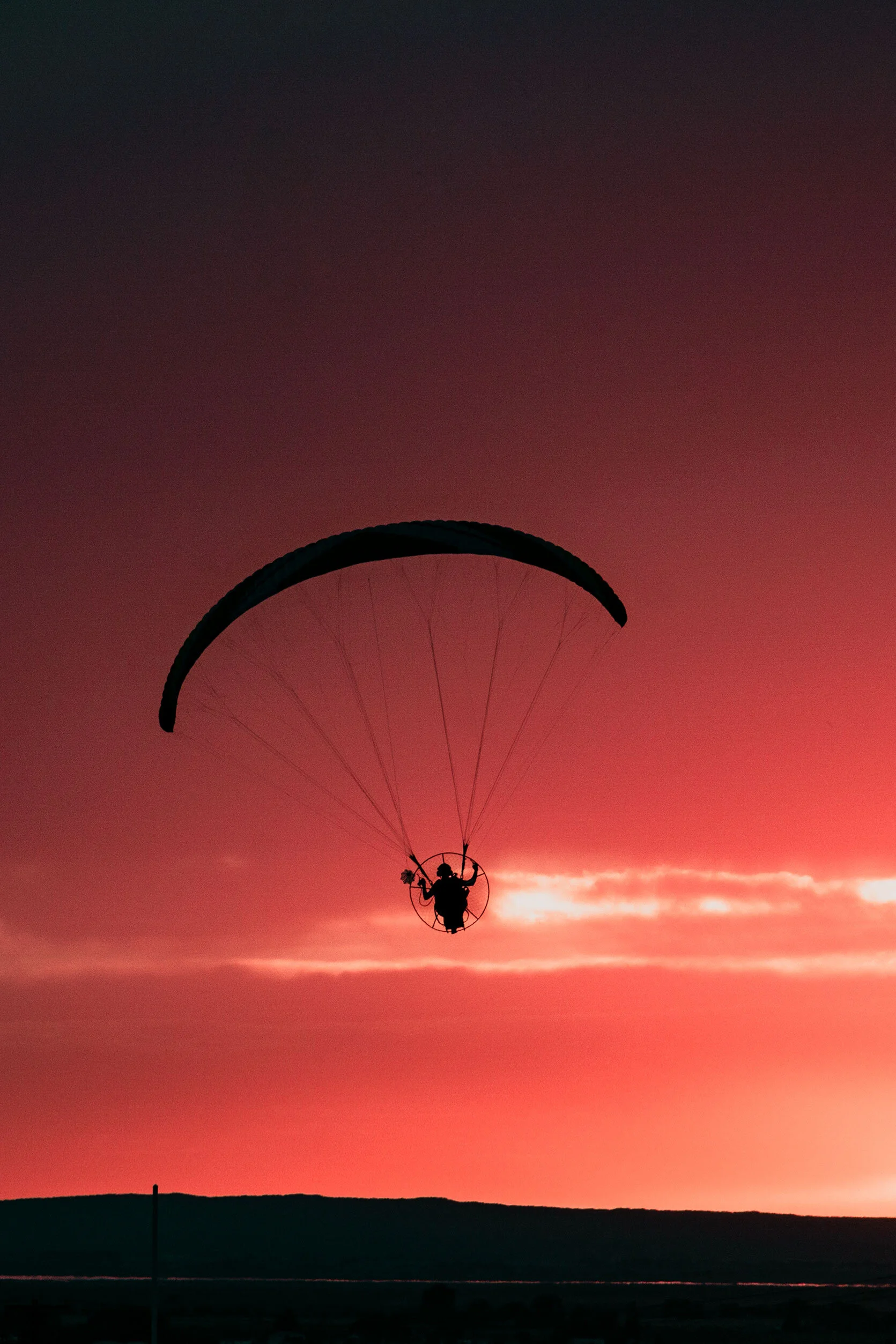 Dark silhouette of guy wearing a motor attached to paraglider flies over black, rolling hills into bright red sunset and wispy colorful clouds. In Pocatello, Idaho.