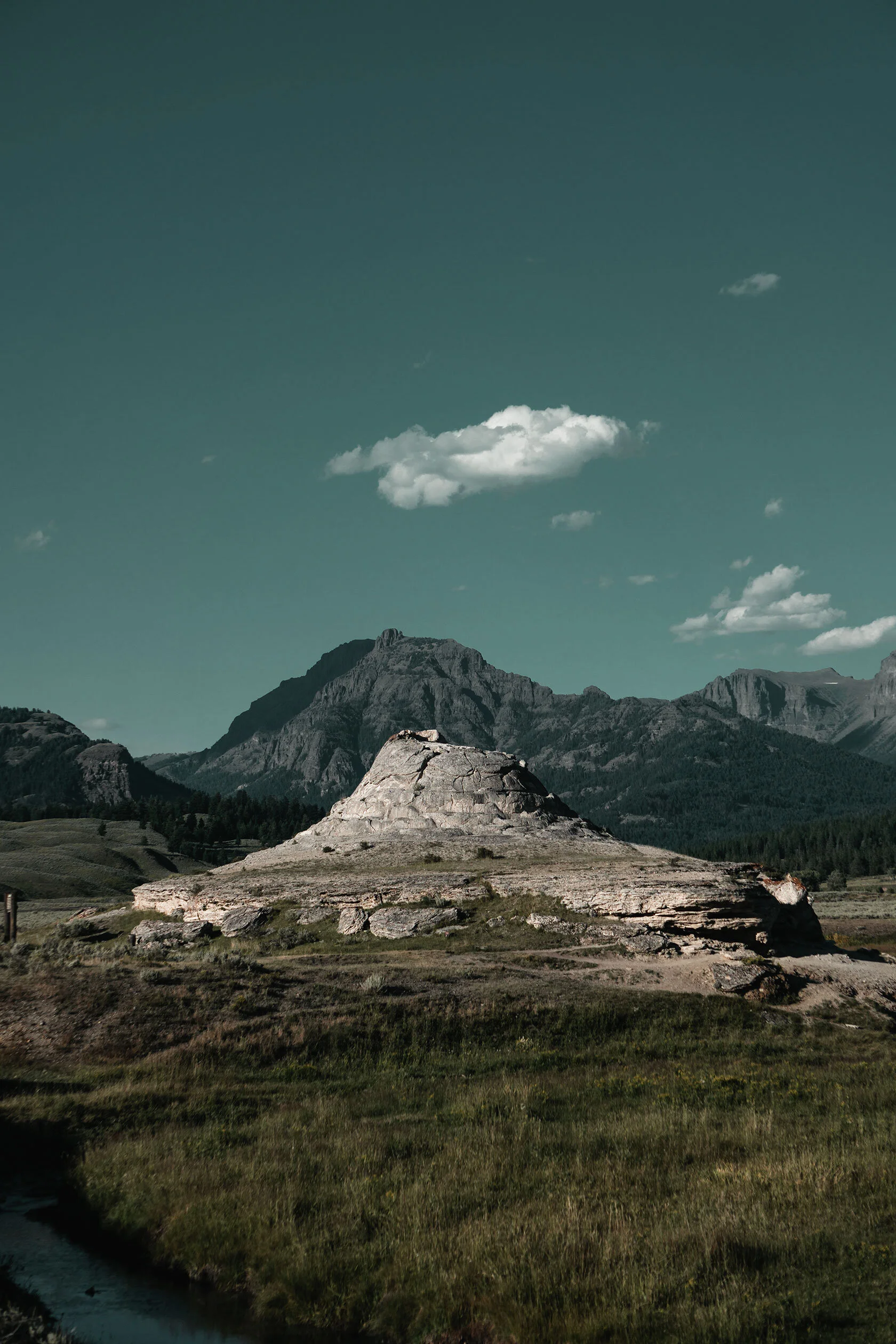 Volcanic formation, resembling a volcano itself, sits inside open plain in Yellowstone volcano under dim grey teal sky doted with white fluffy clouds. Yellowstone National Park, Montana.