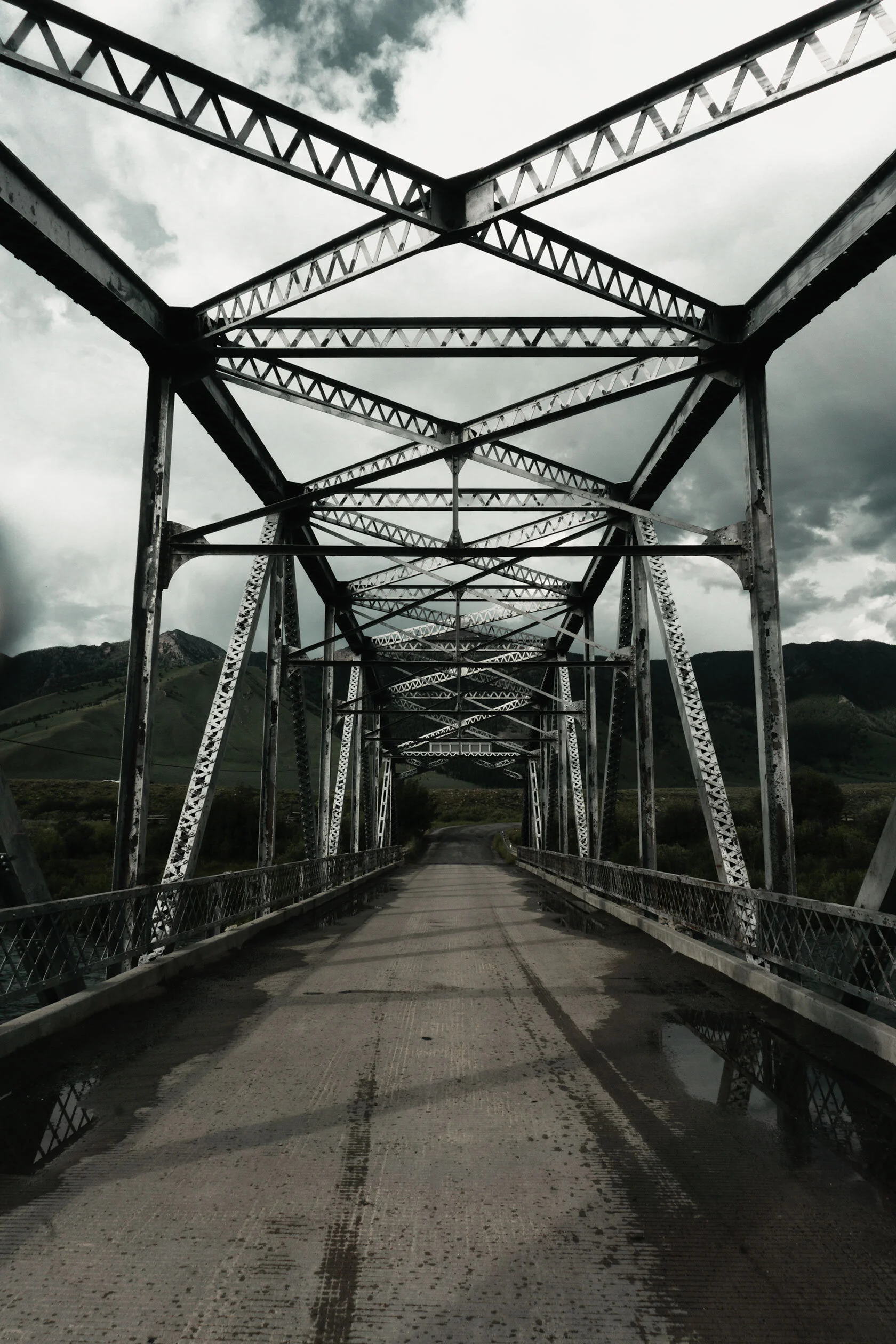 Looking down a rusty metal suspension bridge, the road curves off into green mountains reaching towards an overcast sky somewhere in Montana.