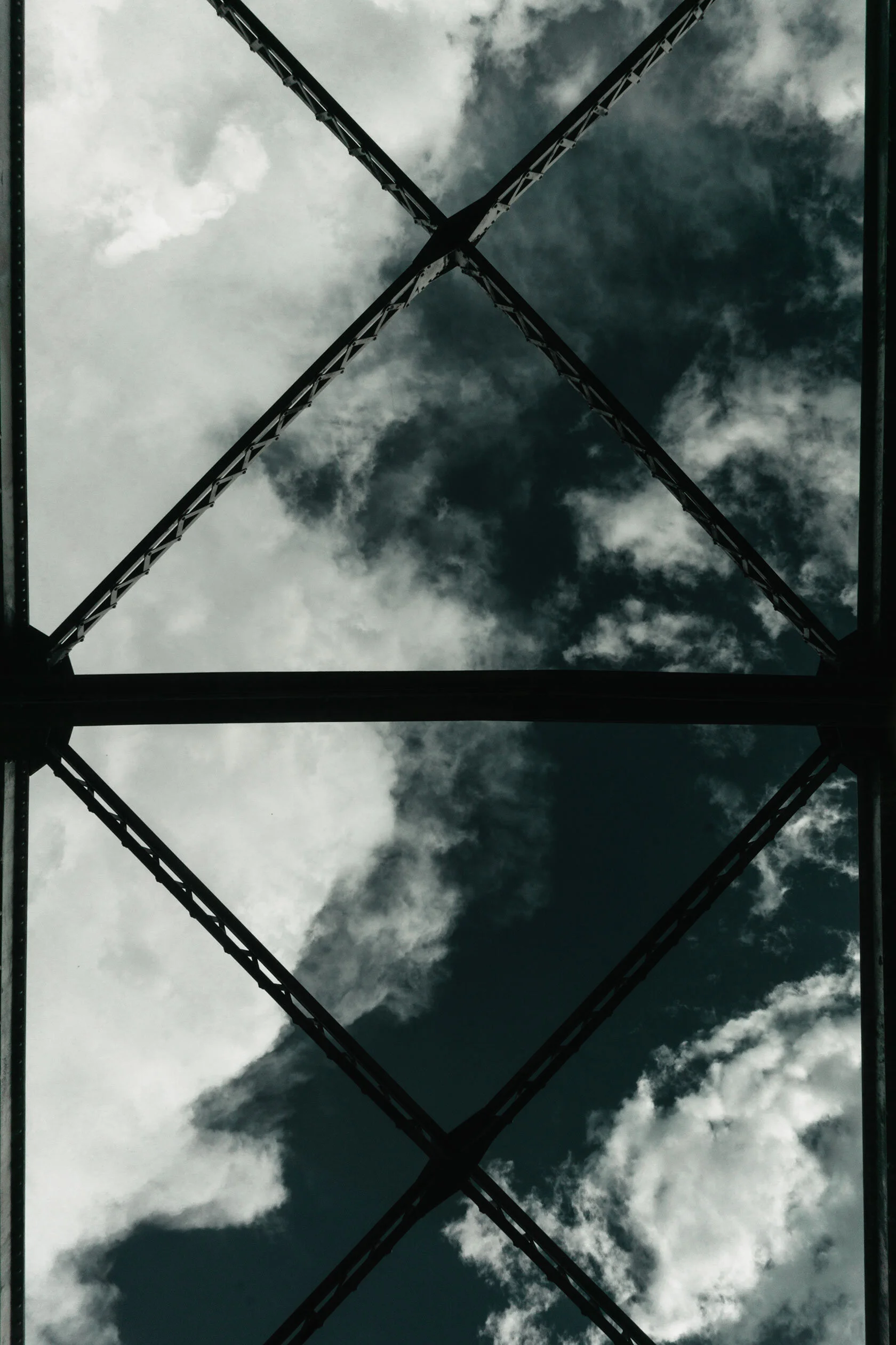 View from below looking straight up at the top of metal suspension bridge in front of dark teal sky peeking through wispy white fluffy clouds after rain. Montana.
