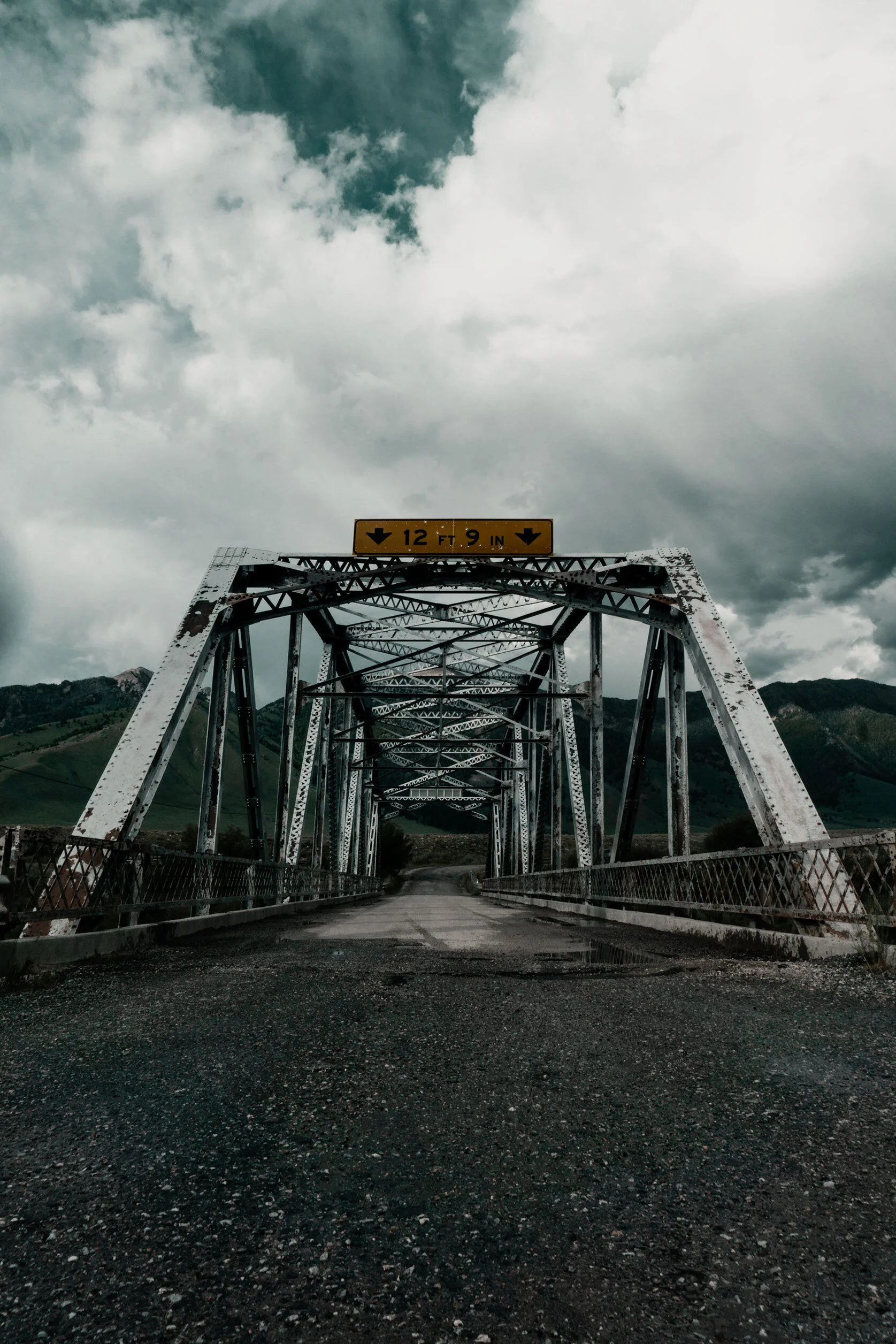 Fluffy white clouds partially obscure dark teal sky over metal suspension bridge from a bug's eye view somewhere in Montana.