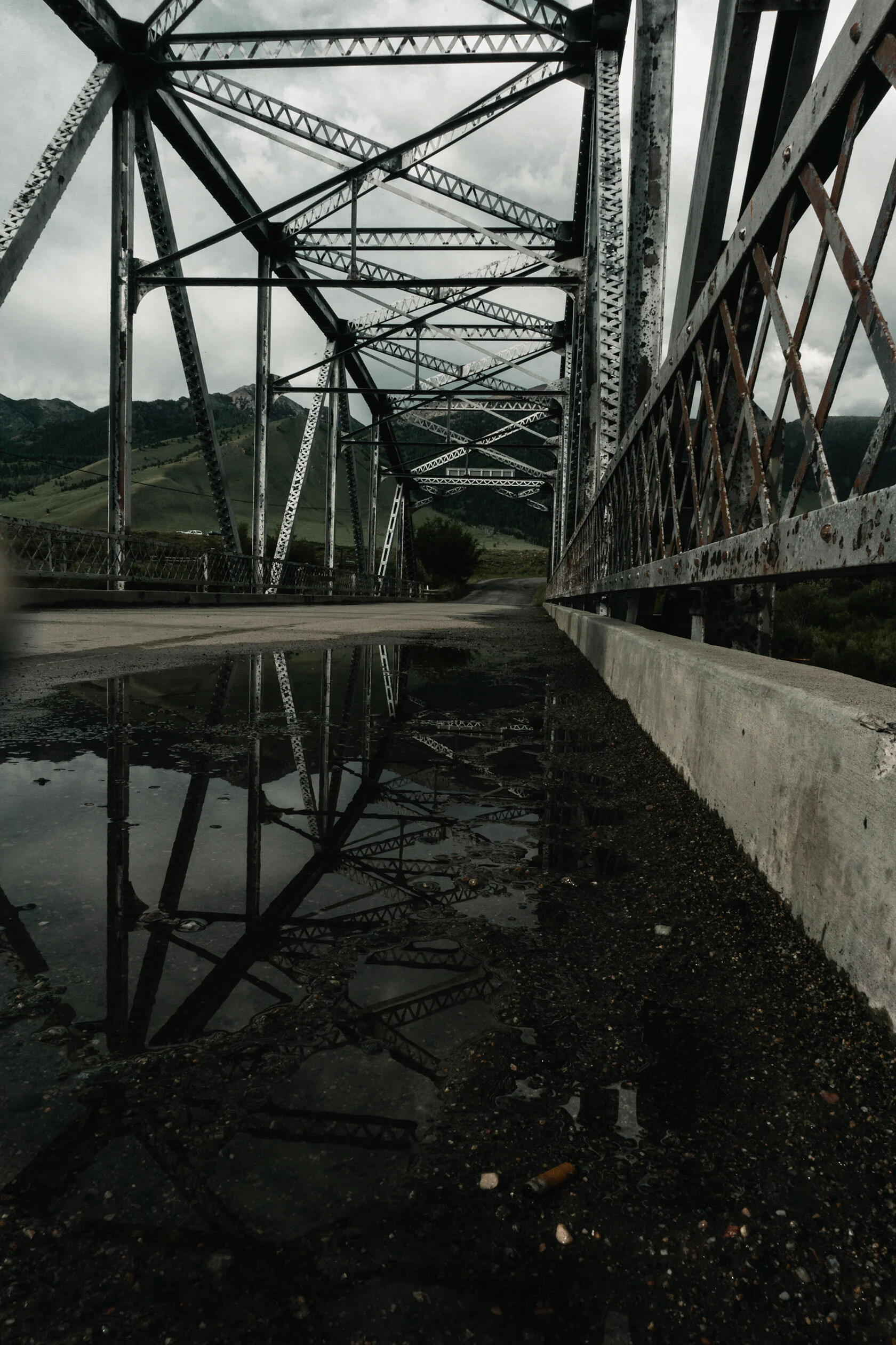 Low angle view looking down rusting metal suspension bridge reflected in a large, still puddle. An old cigarette butt sits near the bottom edge of the frame. Taken in Montana, U.S.A.