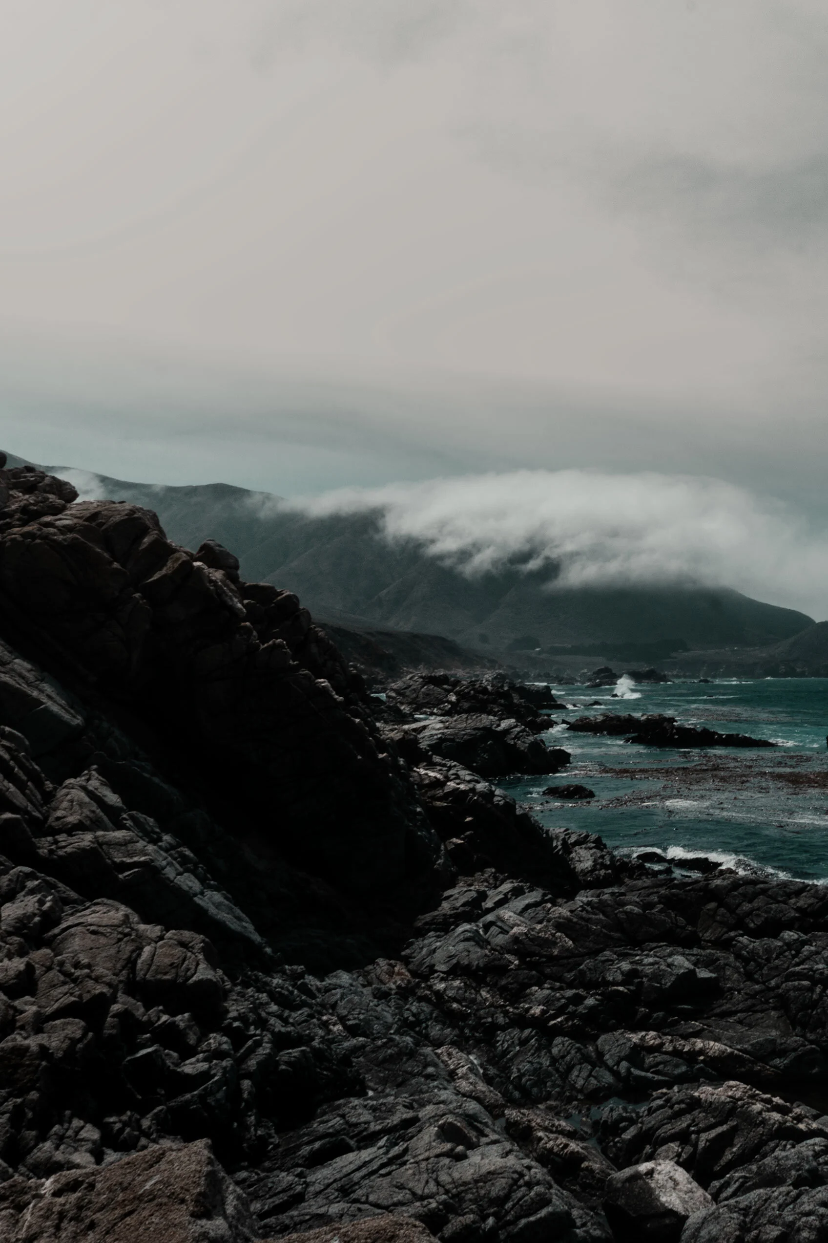 Dark teal sea crashes against black and grey rocks of California coast under cloudy sky.