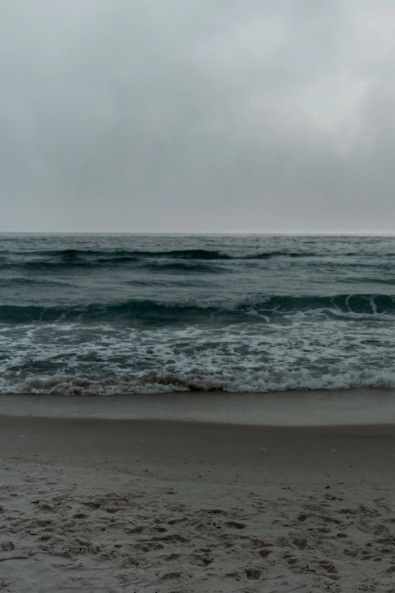 Bug's eye view of Florida beach. Grey sky, dark ocean, teal waves, white sea foam, and light sand form horizontal stripes.